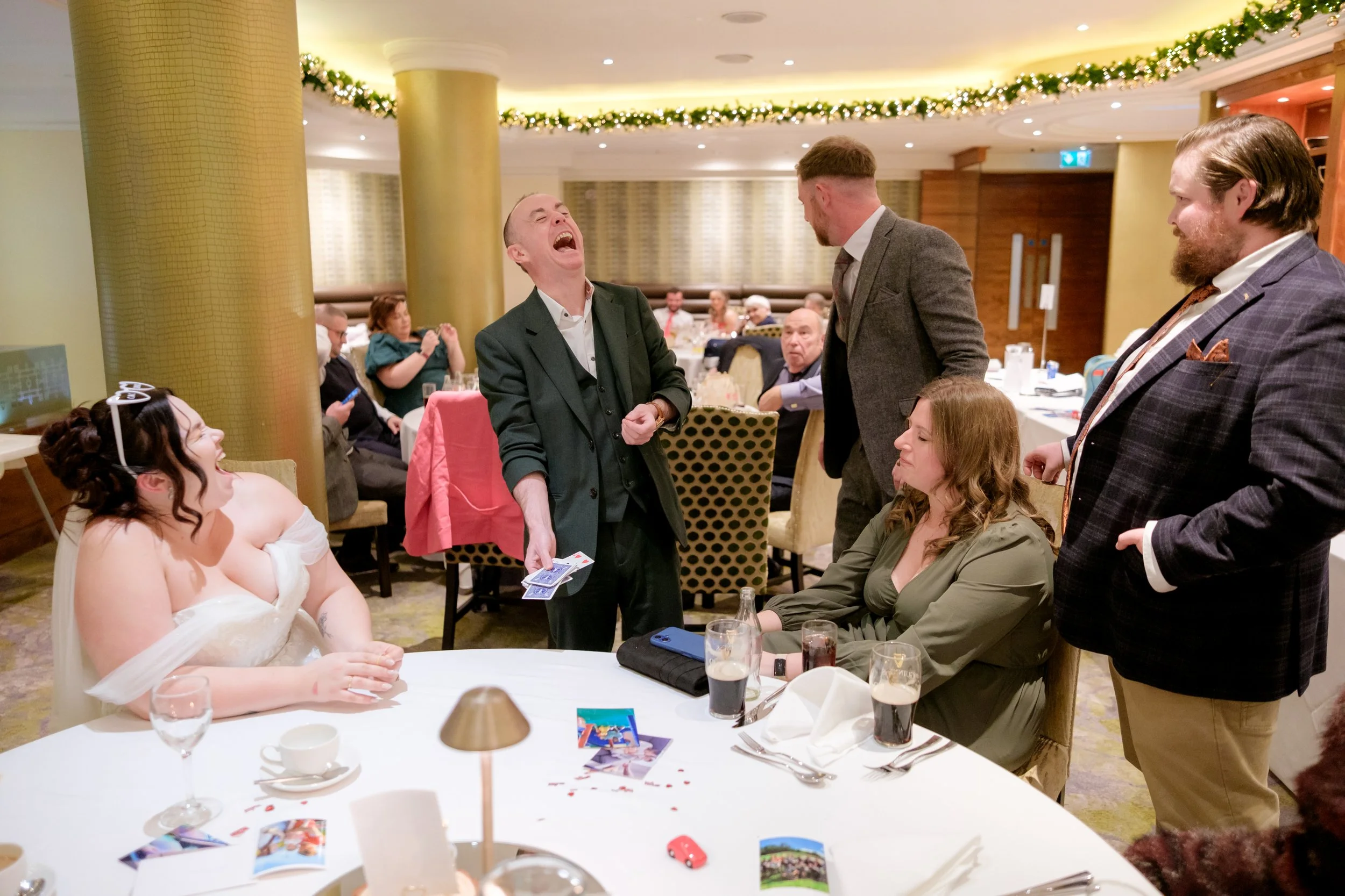 People at a formal event or celebration, with one man holding playing cards and laughing, others watching and interacting, a woman in a white dress sitting at the table, and a decorated banquet hall.