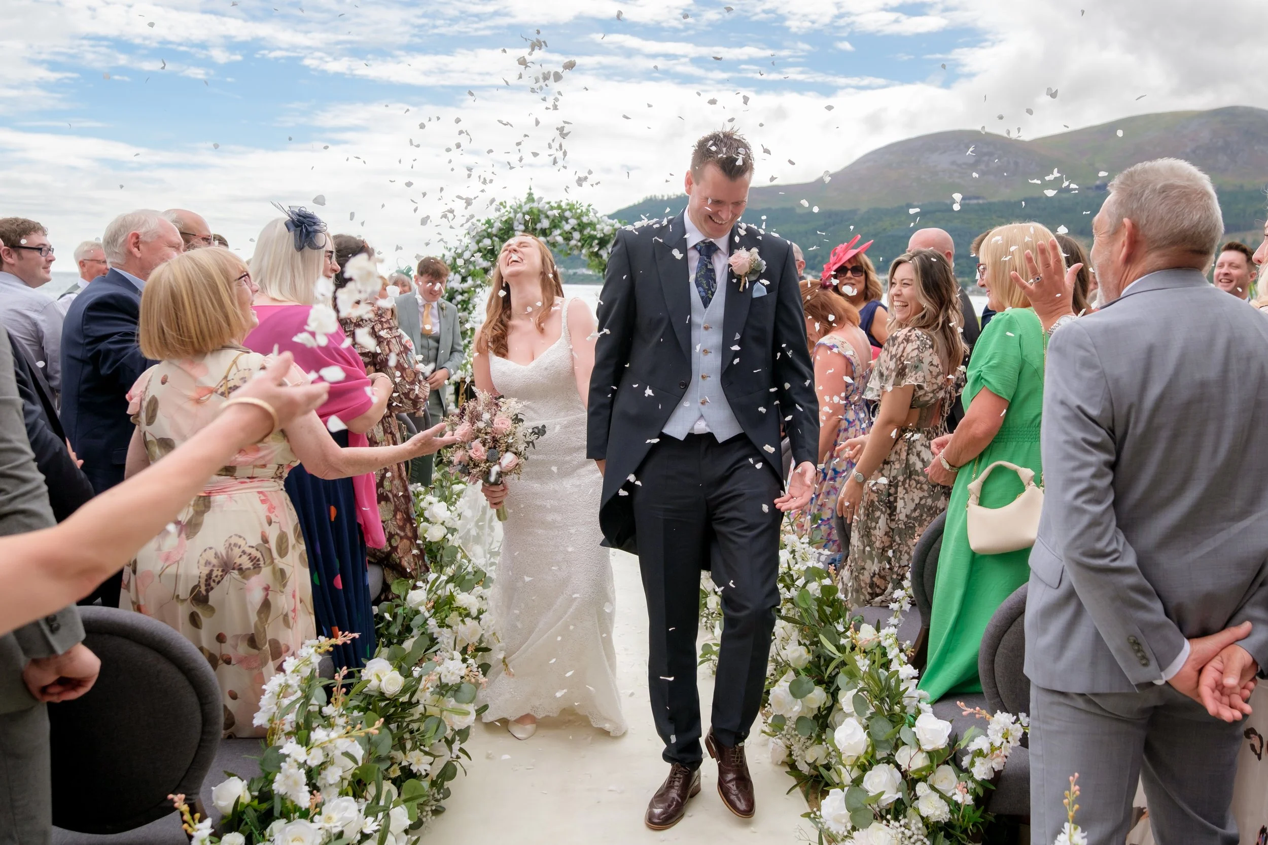 A wedding ceremony with a bride and groom walking down the aisle while guests cheer and throw confetti; outdoor setting with mountains in the background.