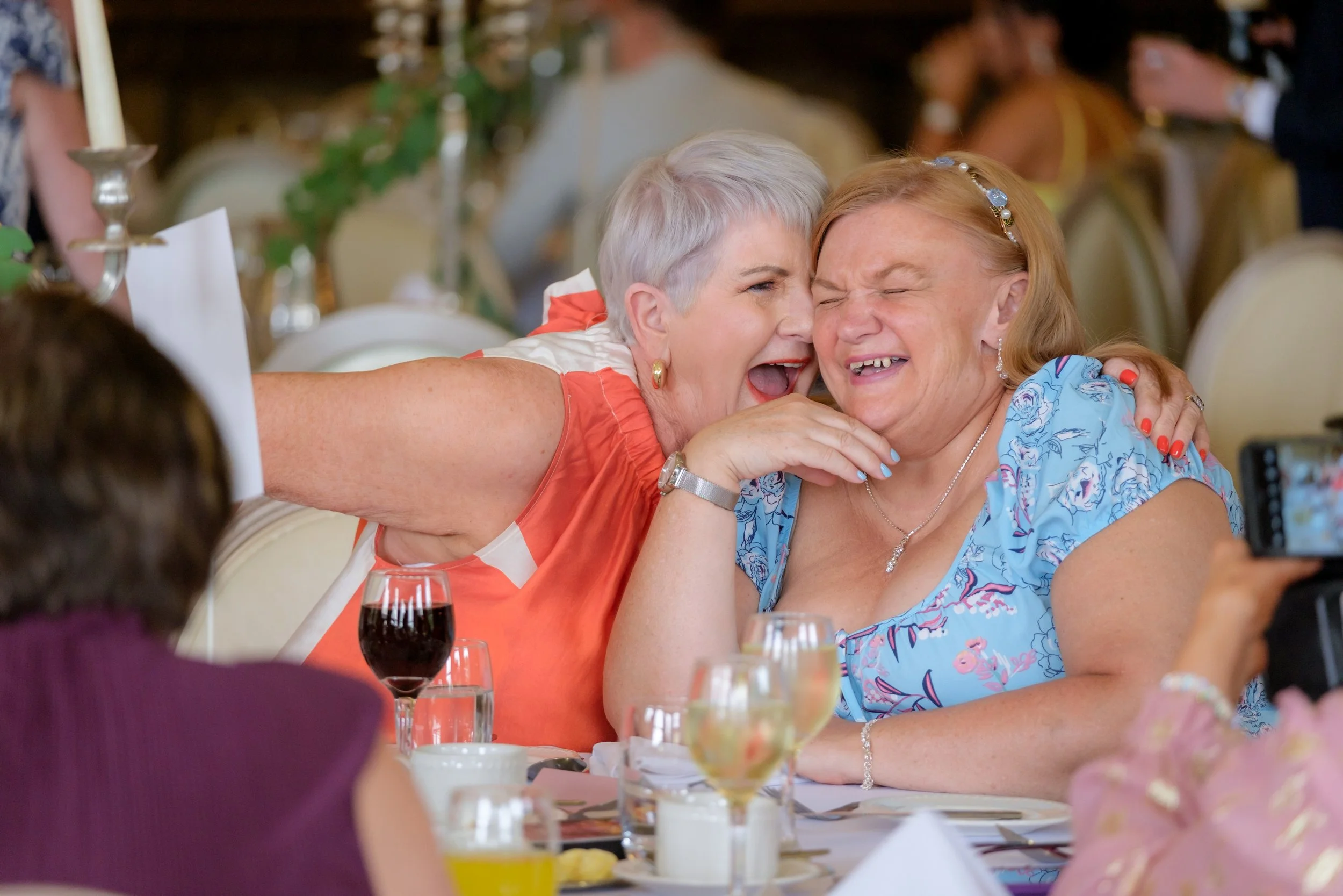 Two women, an older woman with short gray hair in a coral dress and a woman with reddish hair in a light blue dress, are sharing a joyful laugh at a banquet table during a celebration or wedding reception, with glasses of wine and other tableware in 