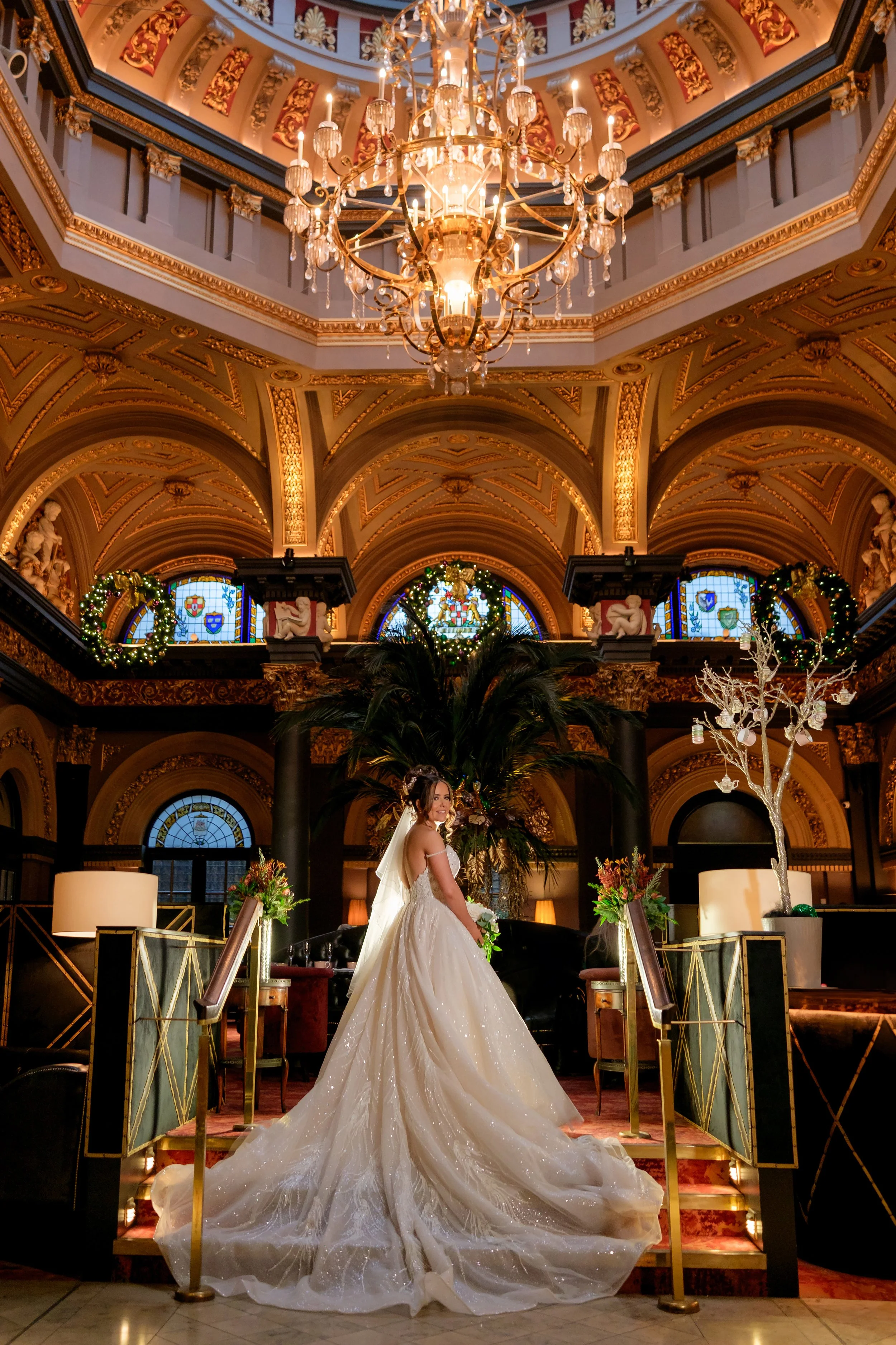 A bride in a white wedding gown standing on a staircase in a luxurious, ornately decorated hall with chandeliers, stained glass windows, and decorated Christmas wreaths and trees.