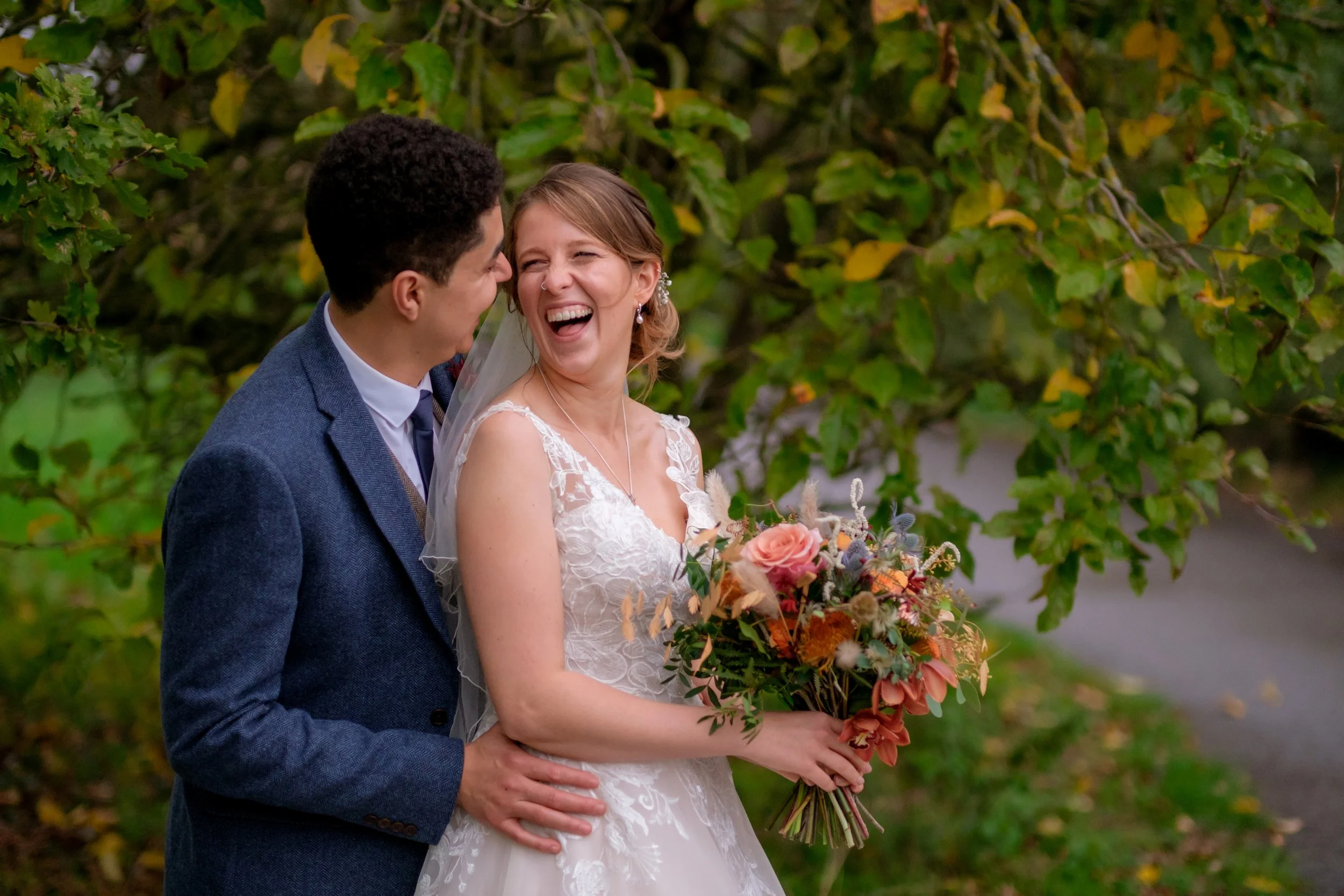 A bride and groom sharing a joyful moment outdoors, with the bride holding a bouquet of flowers and surrounded by green foliage.