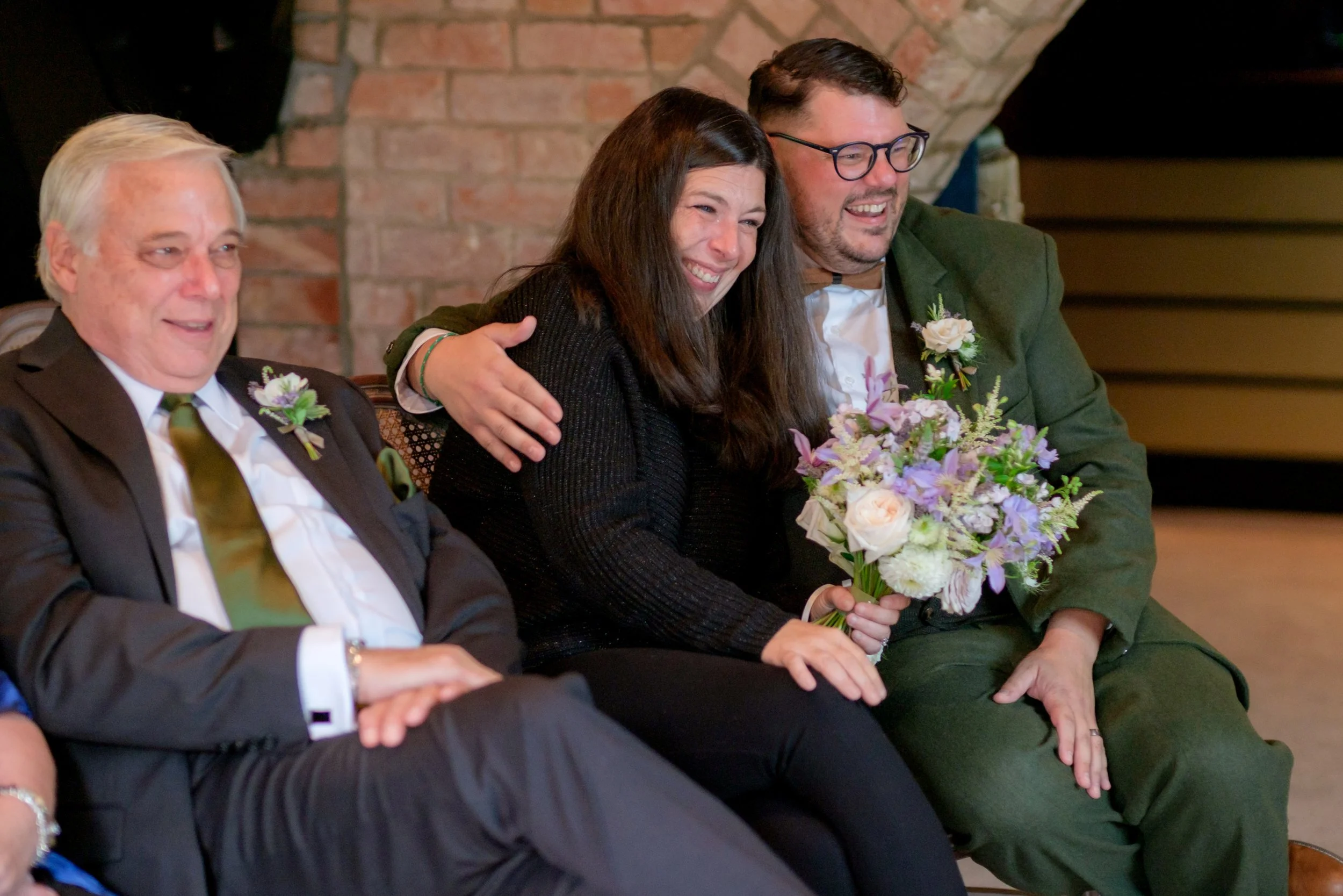 People sitting together at a celebration, with one woman holding a bouquet of flowers, smiling, and wearing dark clothing, while men around her are dressed in suits with boutonnières, in a room with brick walls.