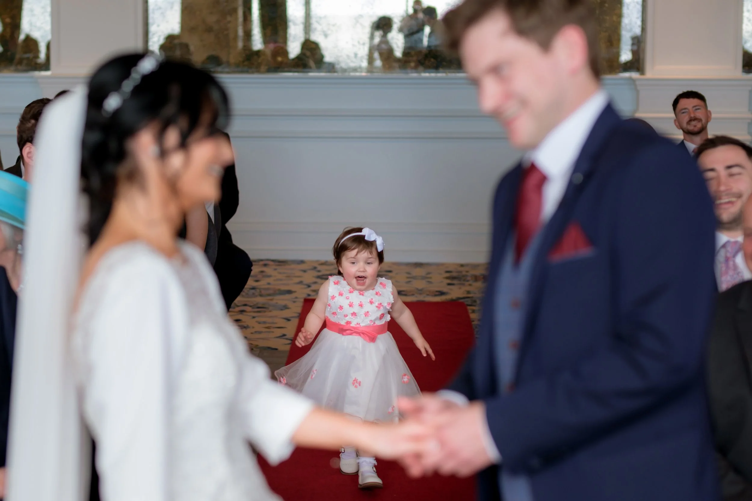A wedding ceremony with the bride and groom holding hands, standing before an officiant, with a young girl in a white dress with pink accents watching and smiling in the background.