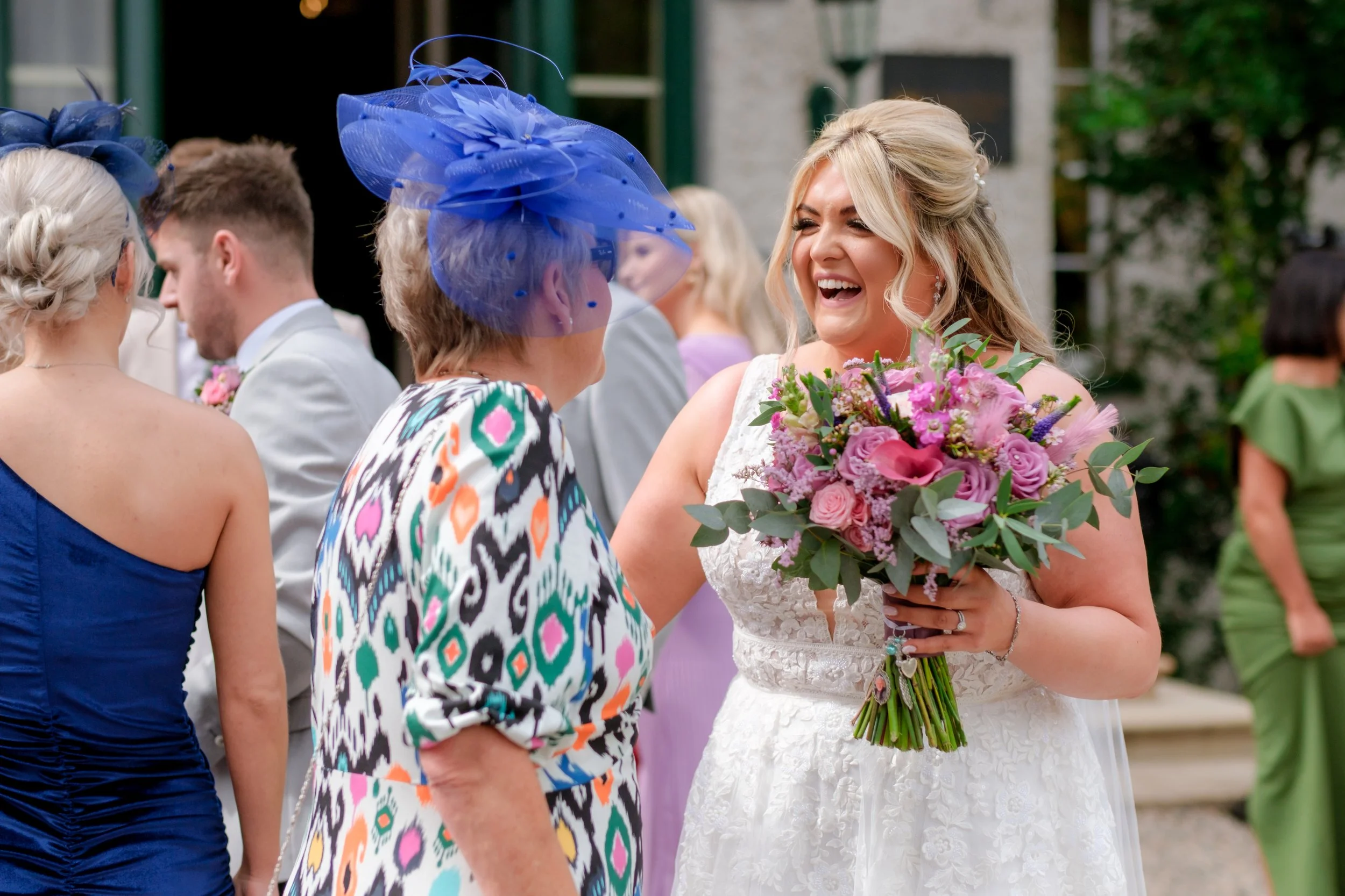 A bride in a white lace wedding dress holding a bouquet of pink and purple flowers, smiling and talking to an older woman with a colorful patterned dress and a large blue hat, at a wedding reception outdoors.