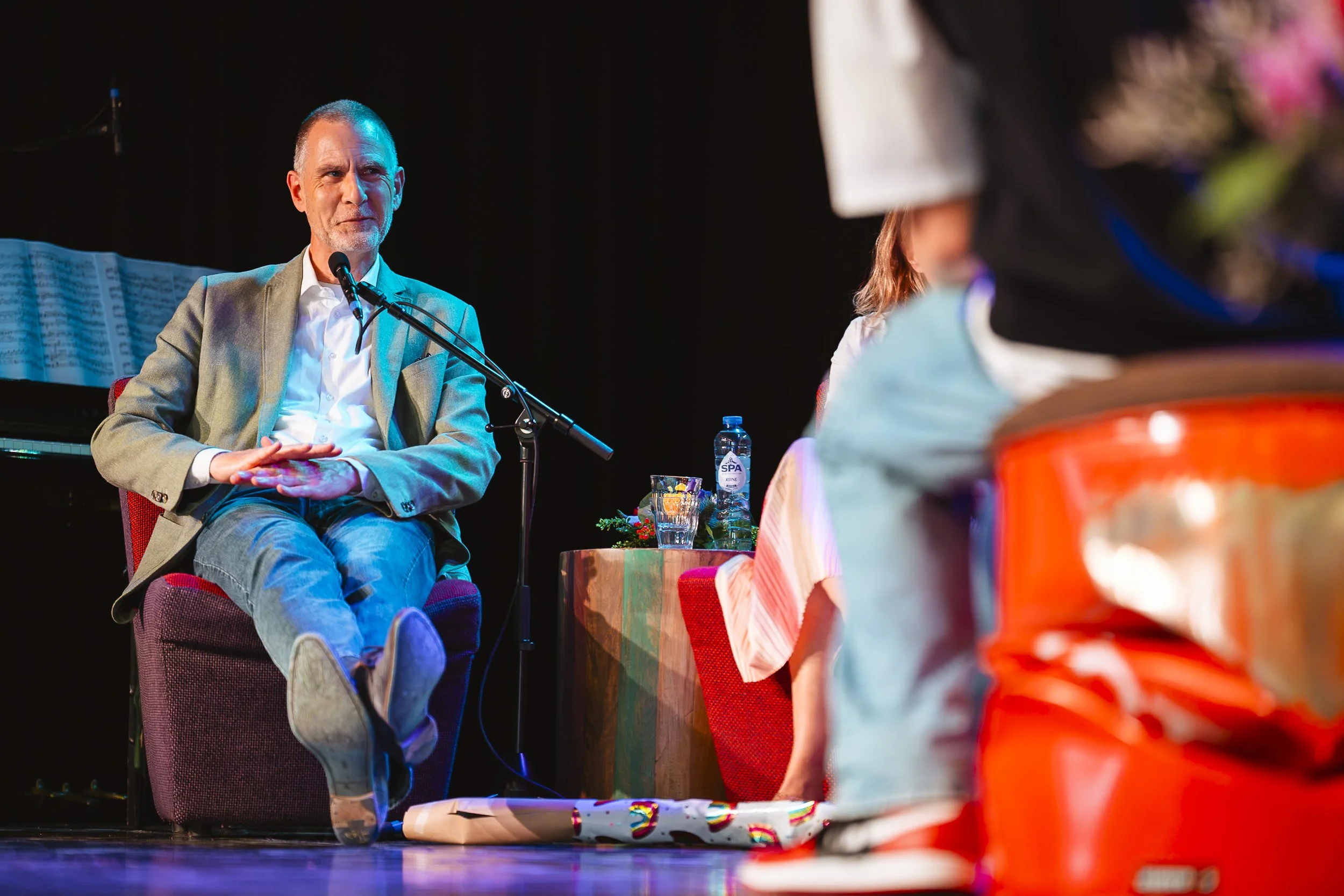 Man zit in een rode fauteuil en spreekt in een microfoon tijdens een bijeenkomst, met een tafel met water en decoraties ernaast.