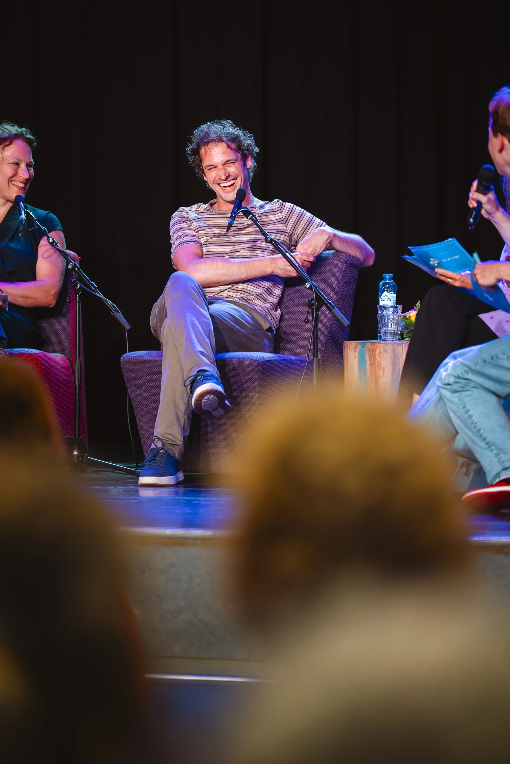 Mensen die op een podium zitten tijdens een interview of discussie, met een man die glimlacht en lacht.