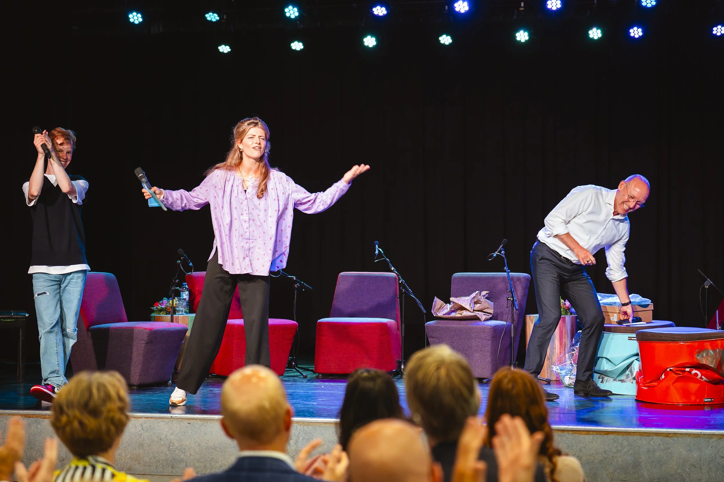 Drie mensen op een podium tijdens een optreed, applaudisserend en met lachende gezichten, met een publiek dat enthousiast klapt.