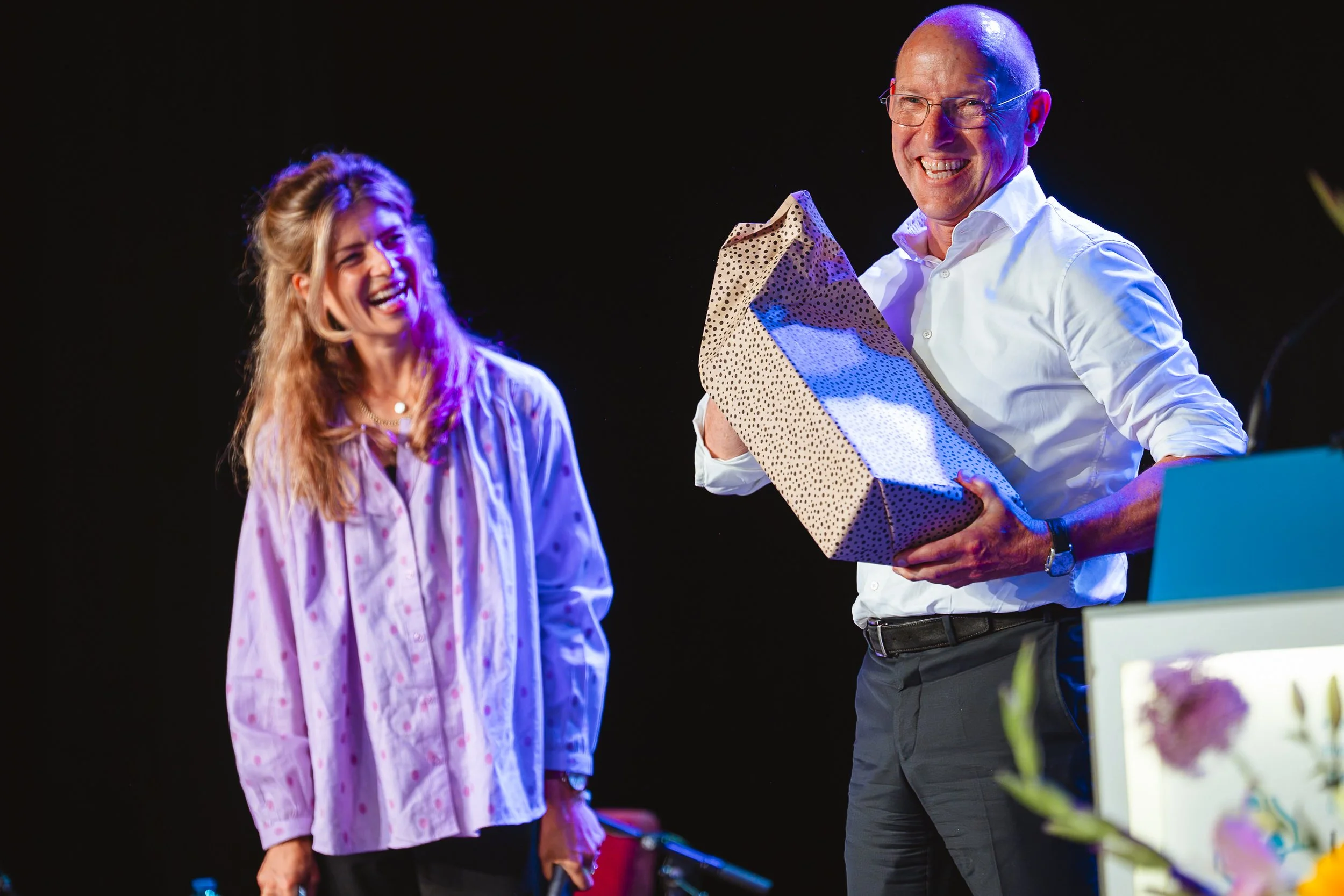 Een lachende vrouw en een lachende man met een cadeau in een donkere setting met kleurrijk licht.