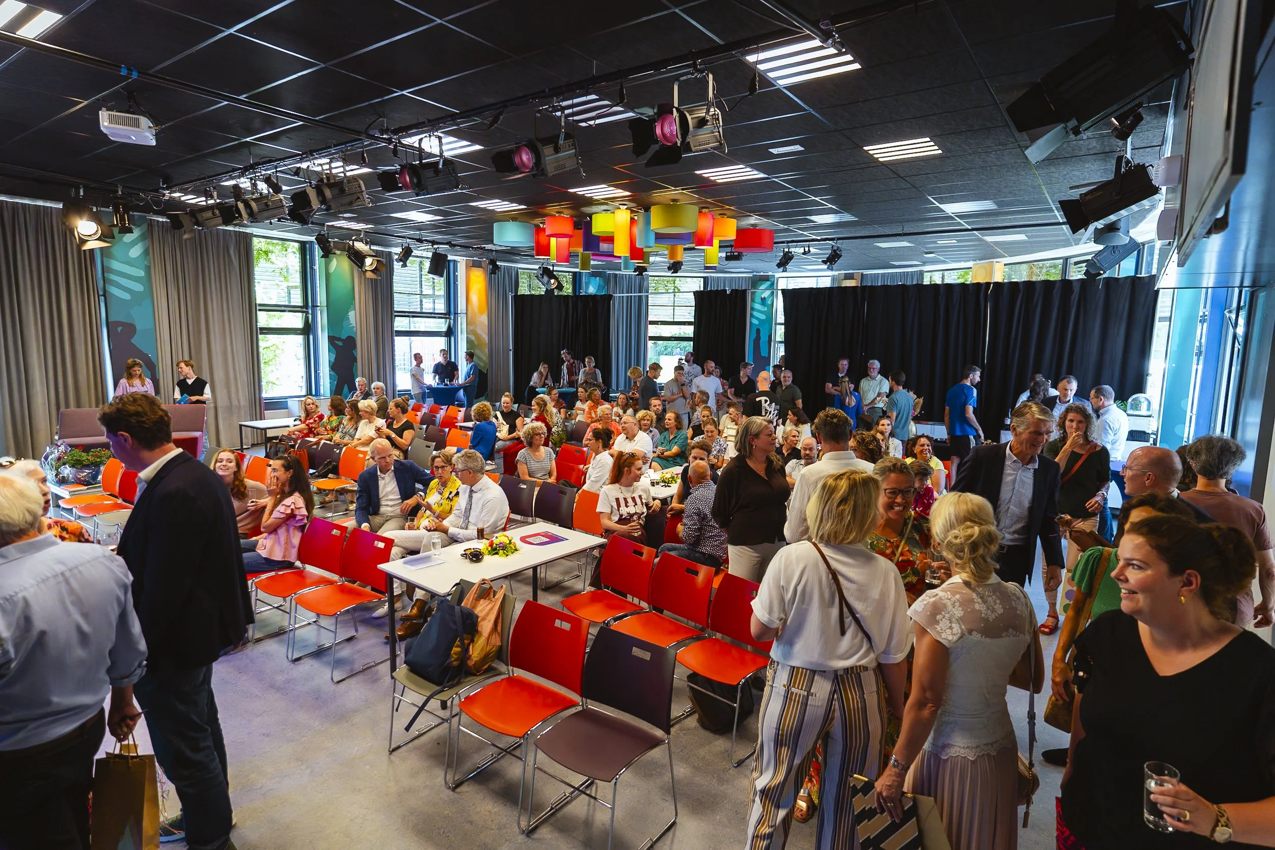 Een drukke zaal met mensen die een evenement bijwonen, met kleurrijke plafondlampen, grote ramen, en een mix van gelopen en zittende gasten.