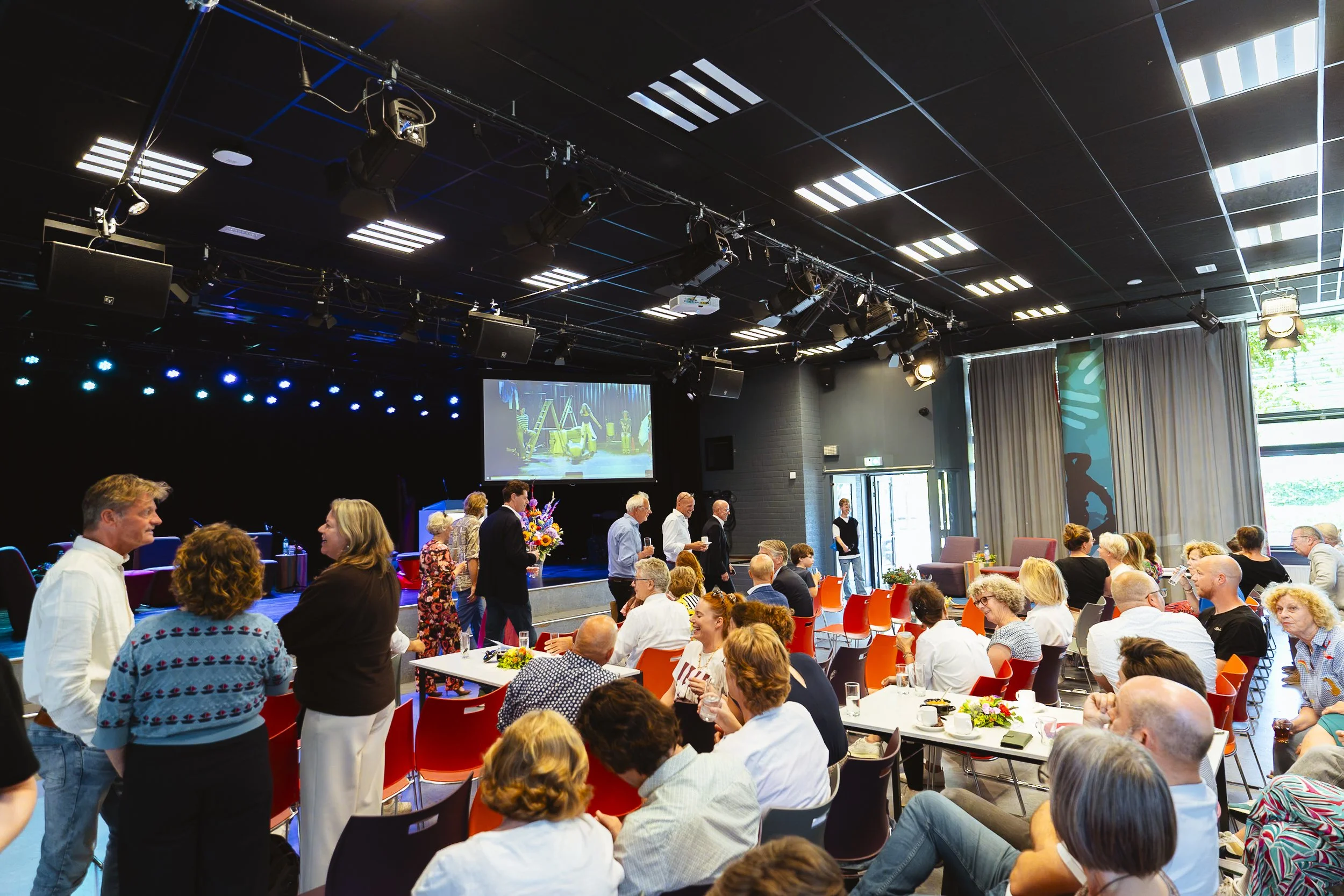 Binnenbeeld van een grote groep mensen die samenkomen in een auditorium of vergaderruimte, met een podium en grote scherm waar een voorstelling of presentatie op wordt getoond.