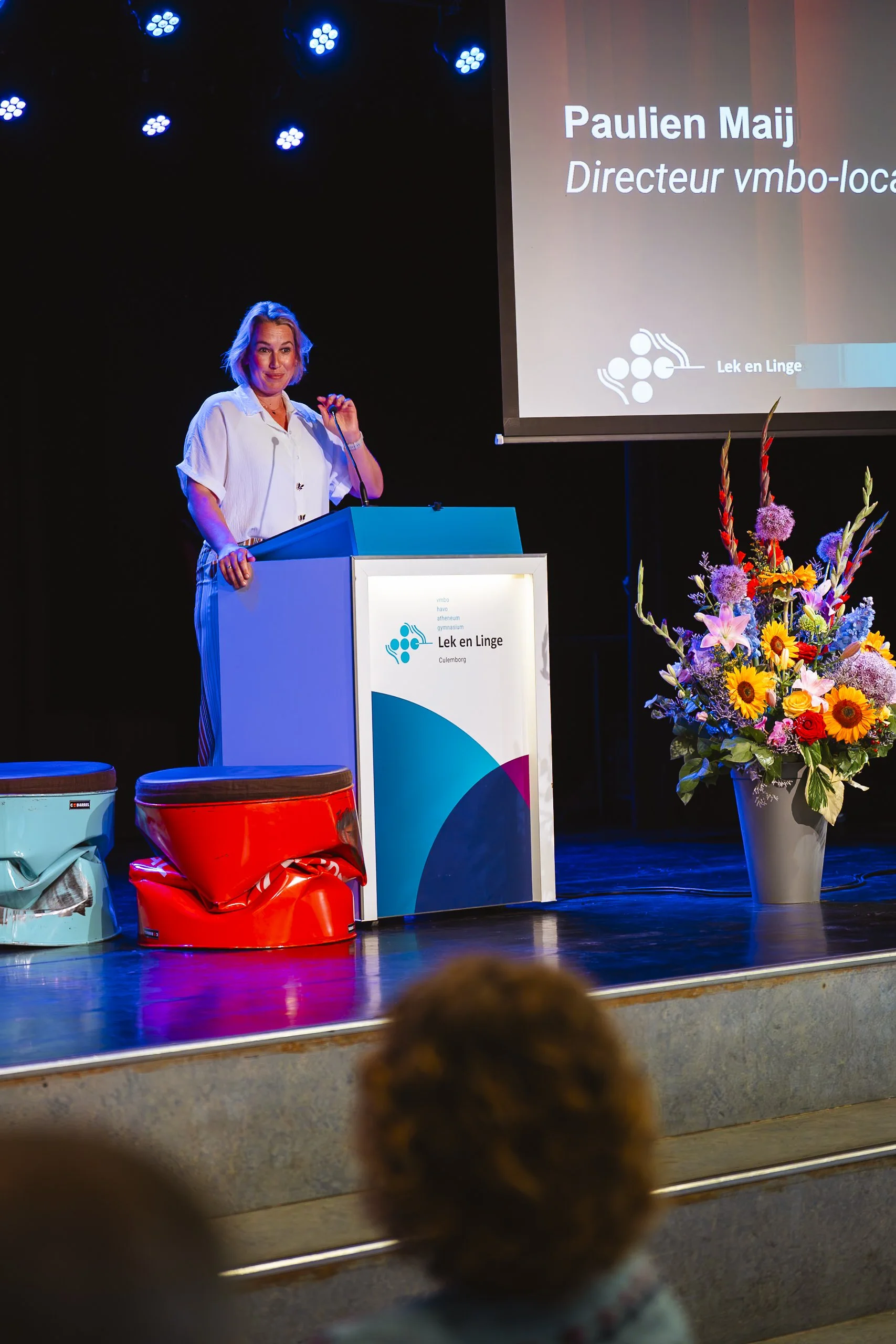 Een vrouw staat achter een rode en een blauwe omwikkelde stoel op een podium, spreekt in een microfoon, met een grote bloemenvaas ernaast. Op het scherm achter haar staat de tekst 'Paulien Maij, Directeur vmbo-loca' en een logo met de woorden 'Lek en