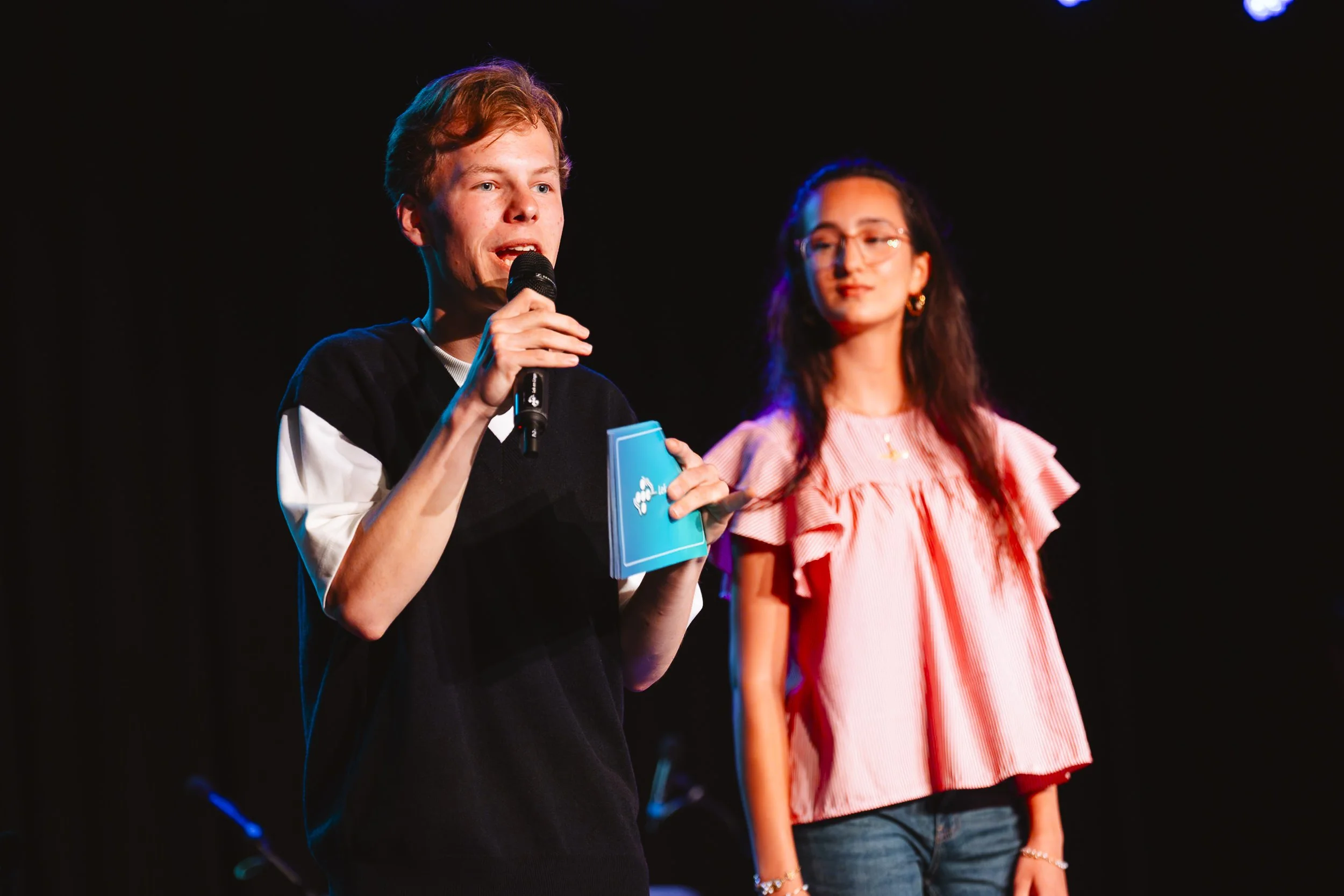 Een jonge man met rood haar die met een microfoon spreekt, terwijl een jonge vrouw met lang haar en bril kijkt en toekijkt tijdens een presentatie op een podium.