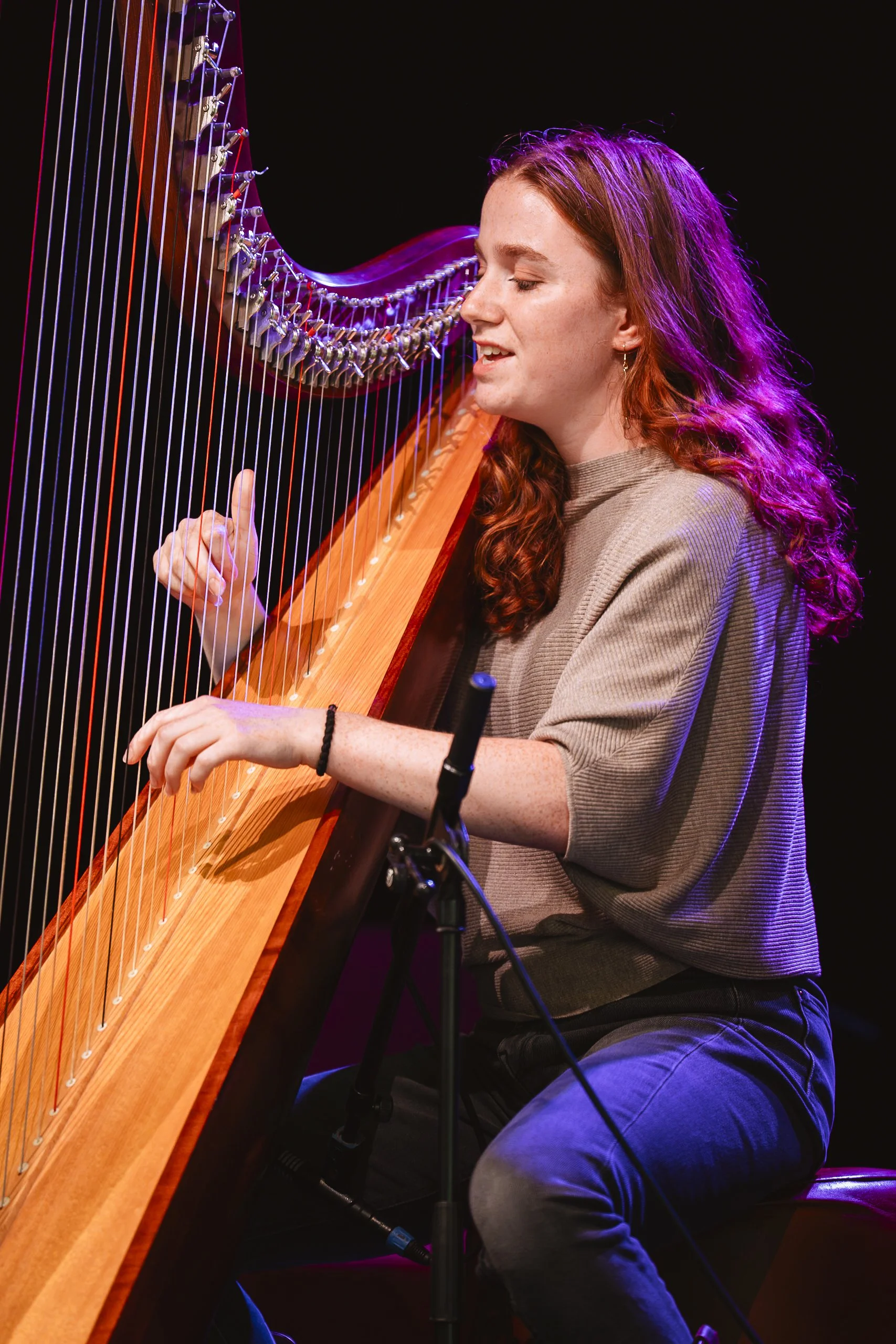 Vrouw speelt harp op podium, met paarse en oranje verlichting, zwarte achtergrond.