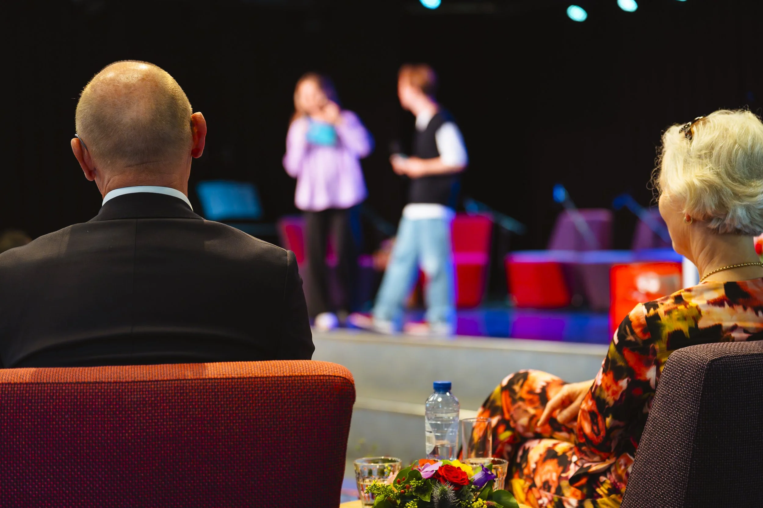 Een man en een vrouw zitten in de zaal en kijken naar een podium waar twee jonge mensen een presentatie geven of optreden, met bloemstuk en drank op de tafel voor hen.