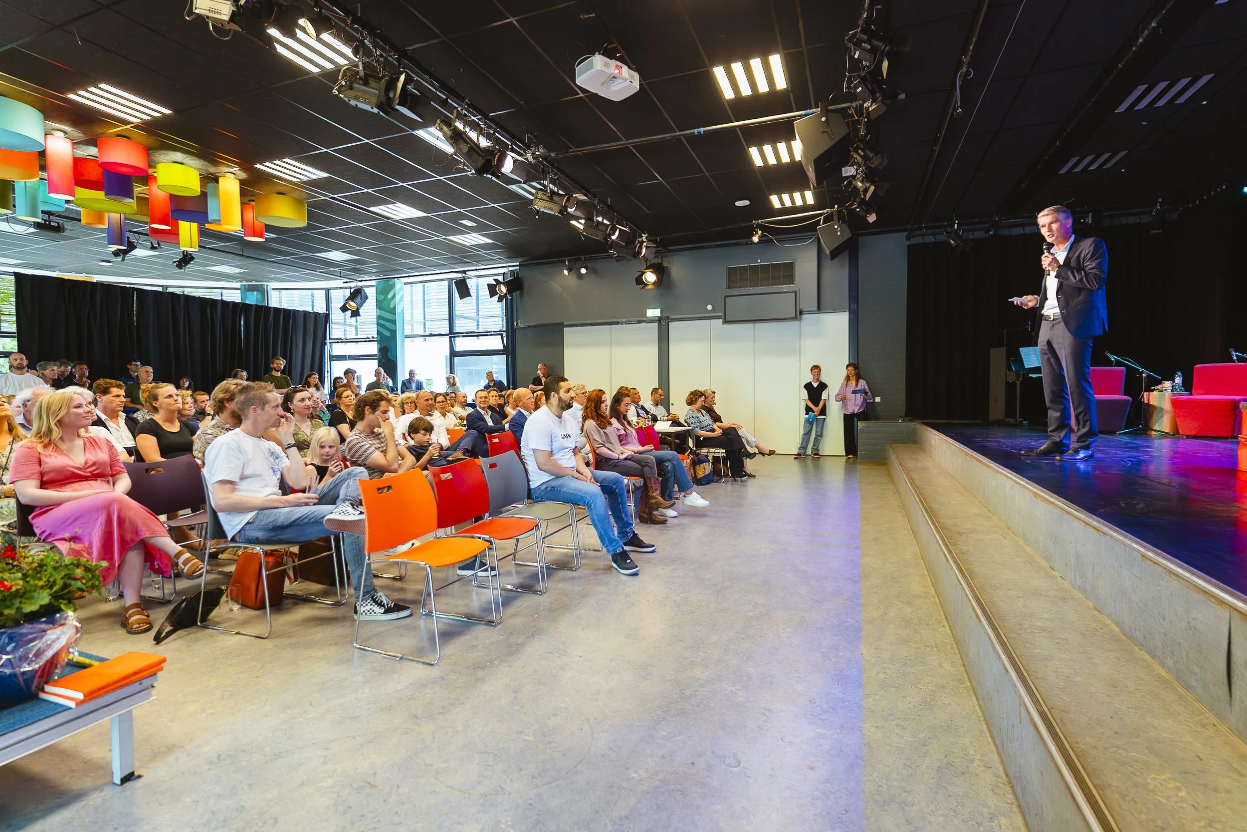 Een auditorium met een podium waar een man in pak met microfoon spreekt voor een publiek dat op stoelen zit, met kleurrijke decoraties en zwart gordijn op de achtergrond.