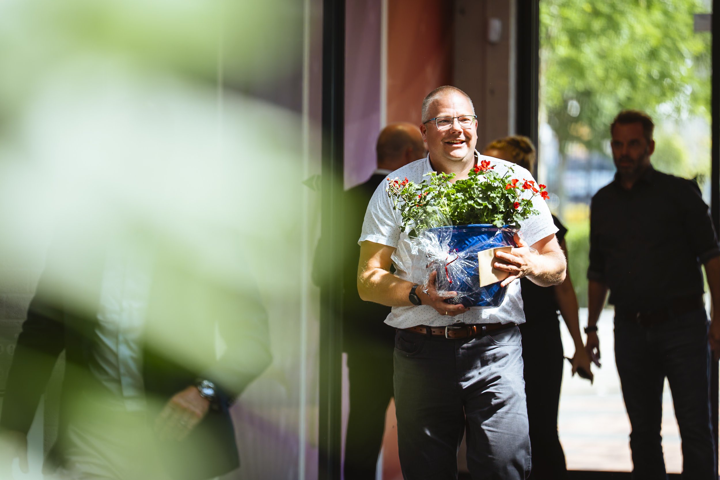 Man die een bloemstuk met rode bloemen vasthoudt en glimlacht, in een inkomhal met anderen op de achtergrond, buiten licht zichtbaar door grote ramen.