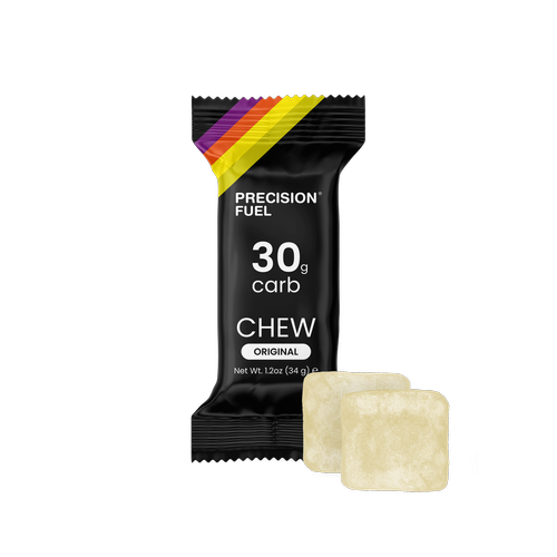 A black packet labeled 'Precision Fuel 30g carb chew original' with two light-colored chew candies in front of it on a black background.