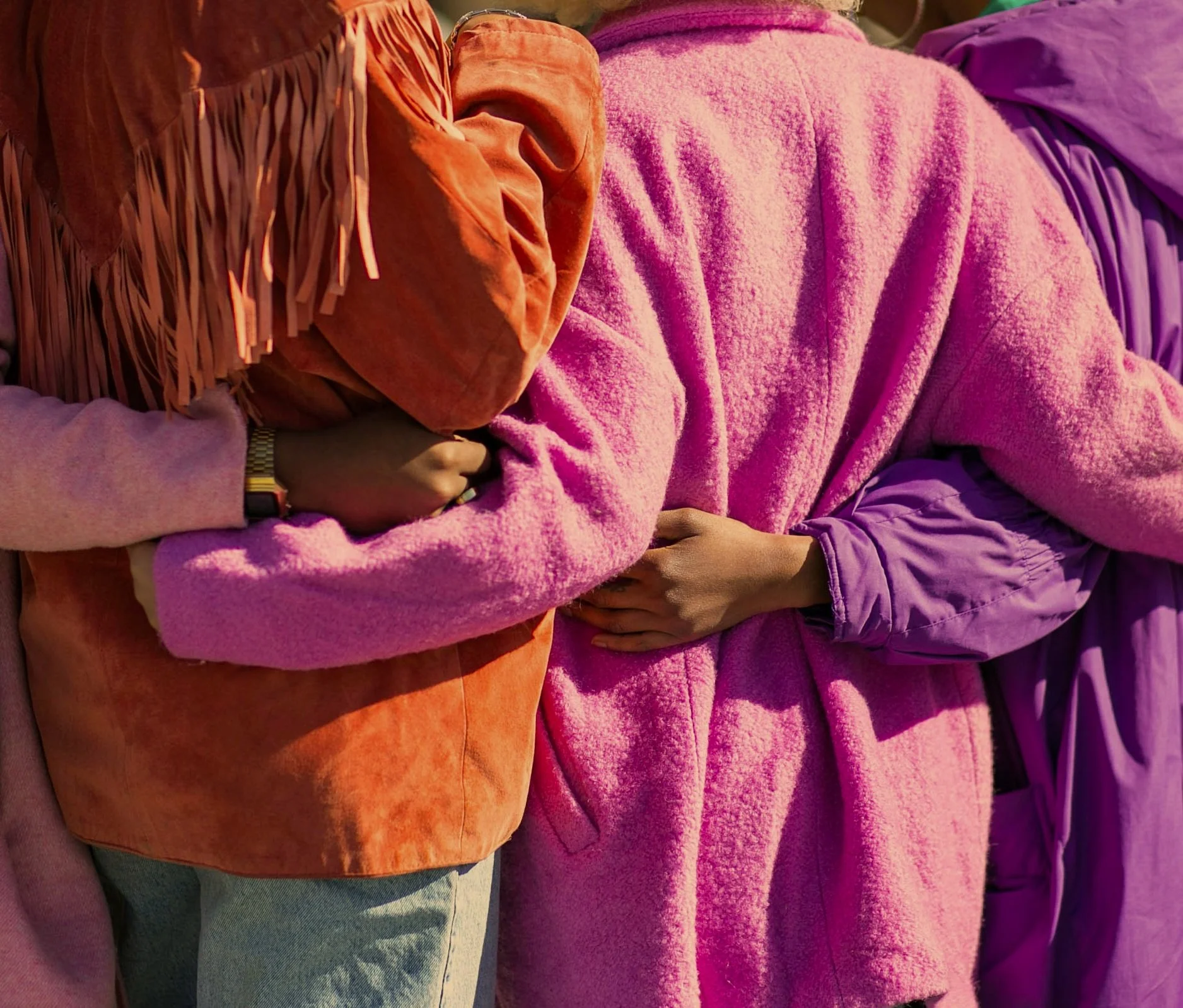 Close-up of four people standing side by side, wearing colorful jackets and sweaters in shades of orange, pink, purple, and lavender, with their arms crossed or holding hands.
