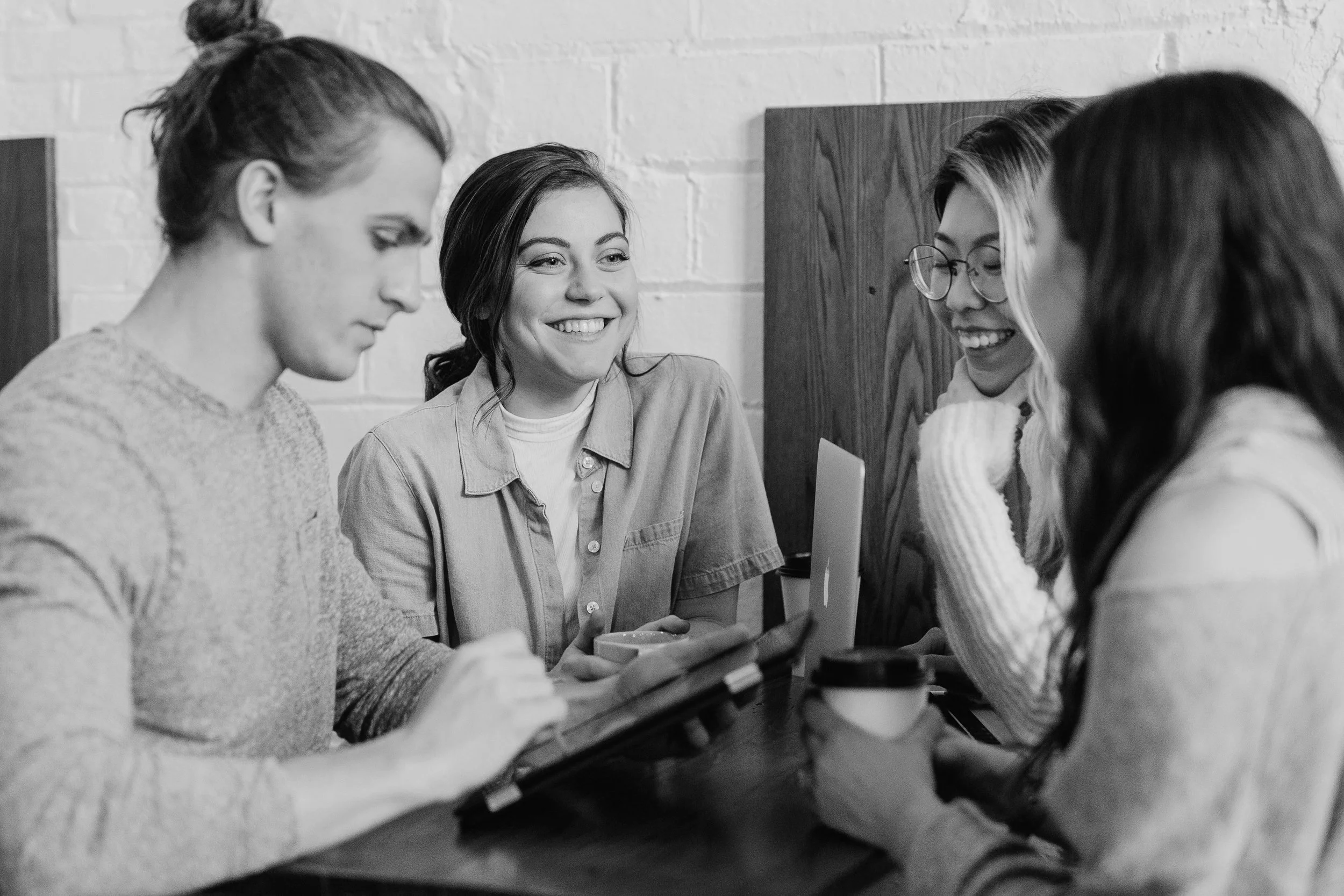 Five women sitting around a table, engaging in a conversation, with one woman showing something on her tablet, smiling and enjoying each other's company.