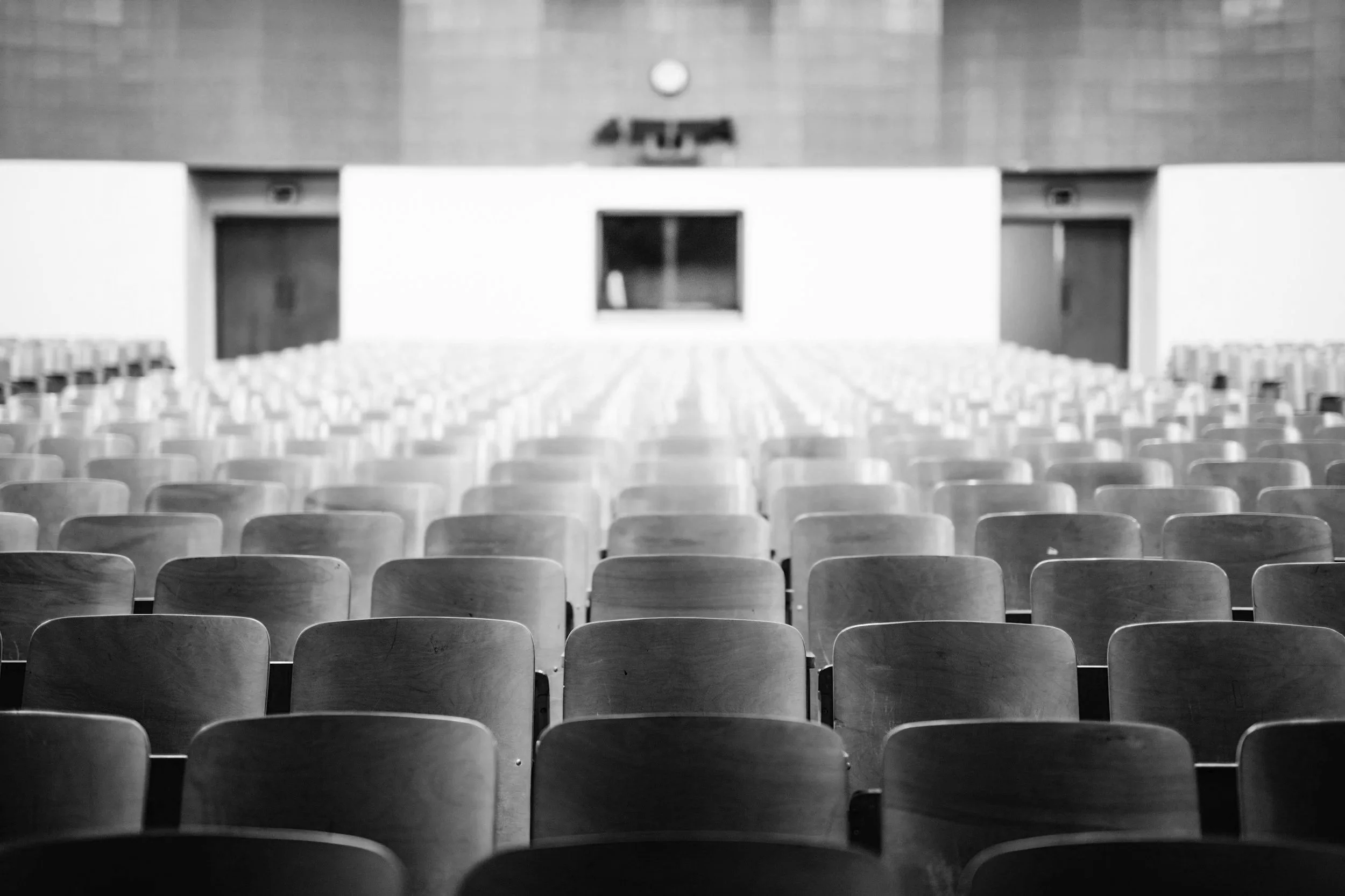 Empty auditorium with rows of chairs facing a stage or screen, with a clock on the wall.