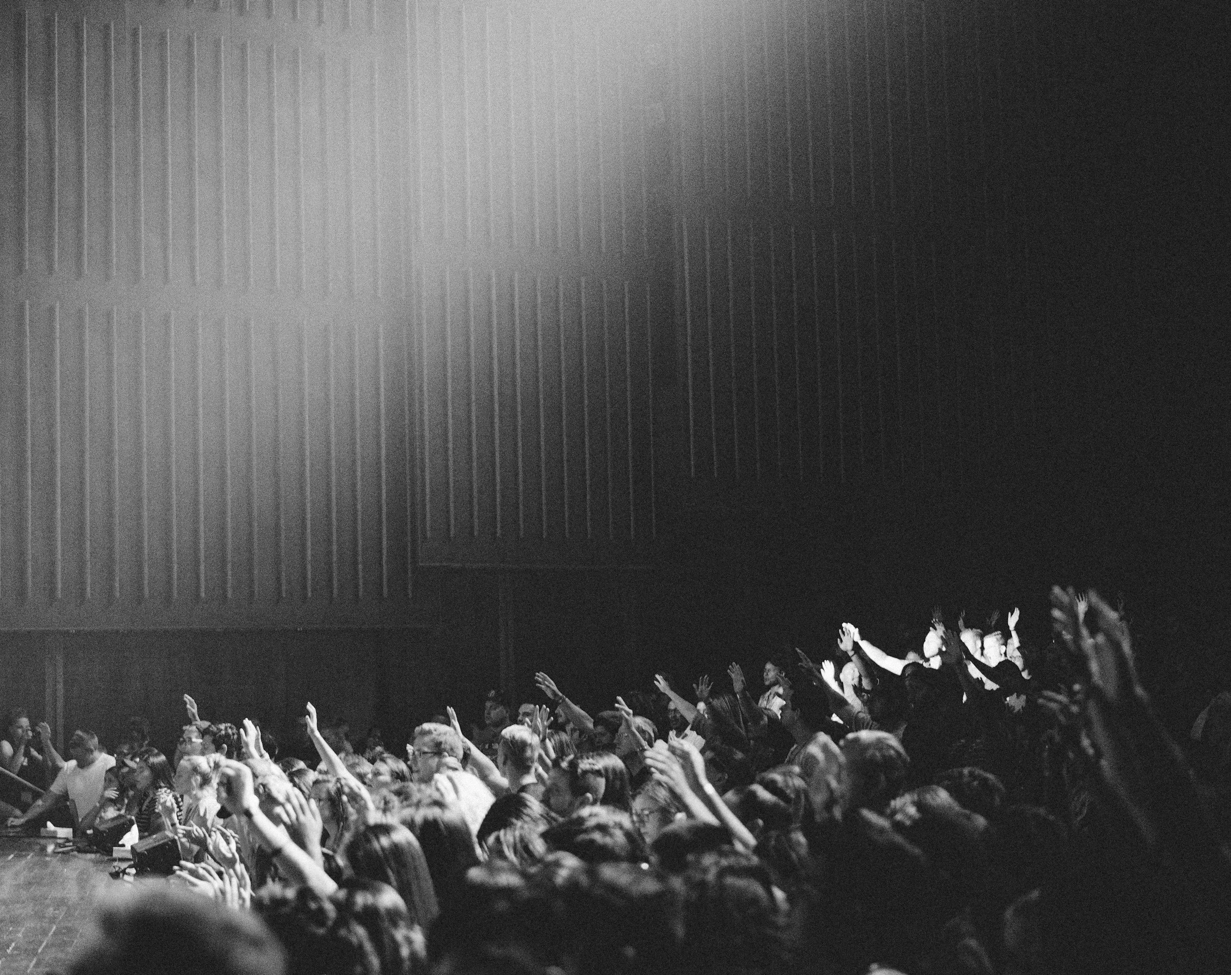 A large audience in a dark auditorium with many people raising their hands, watching a presentation or performance on stage, which is not visible in the image.