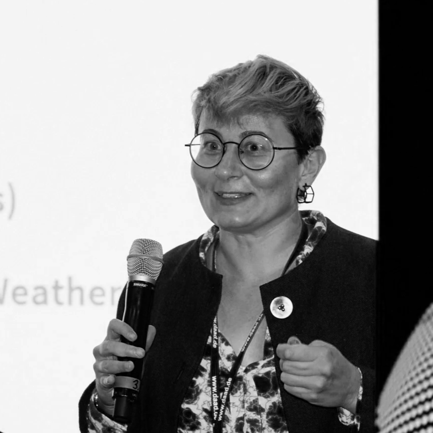 A woman with short hair and glasses speaking into a microphone, wearing a dark jacket and patterned blouse, at an event or presentation.