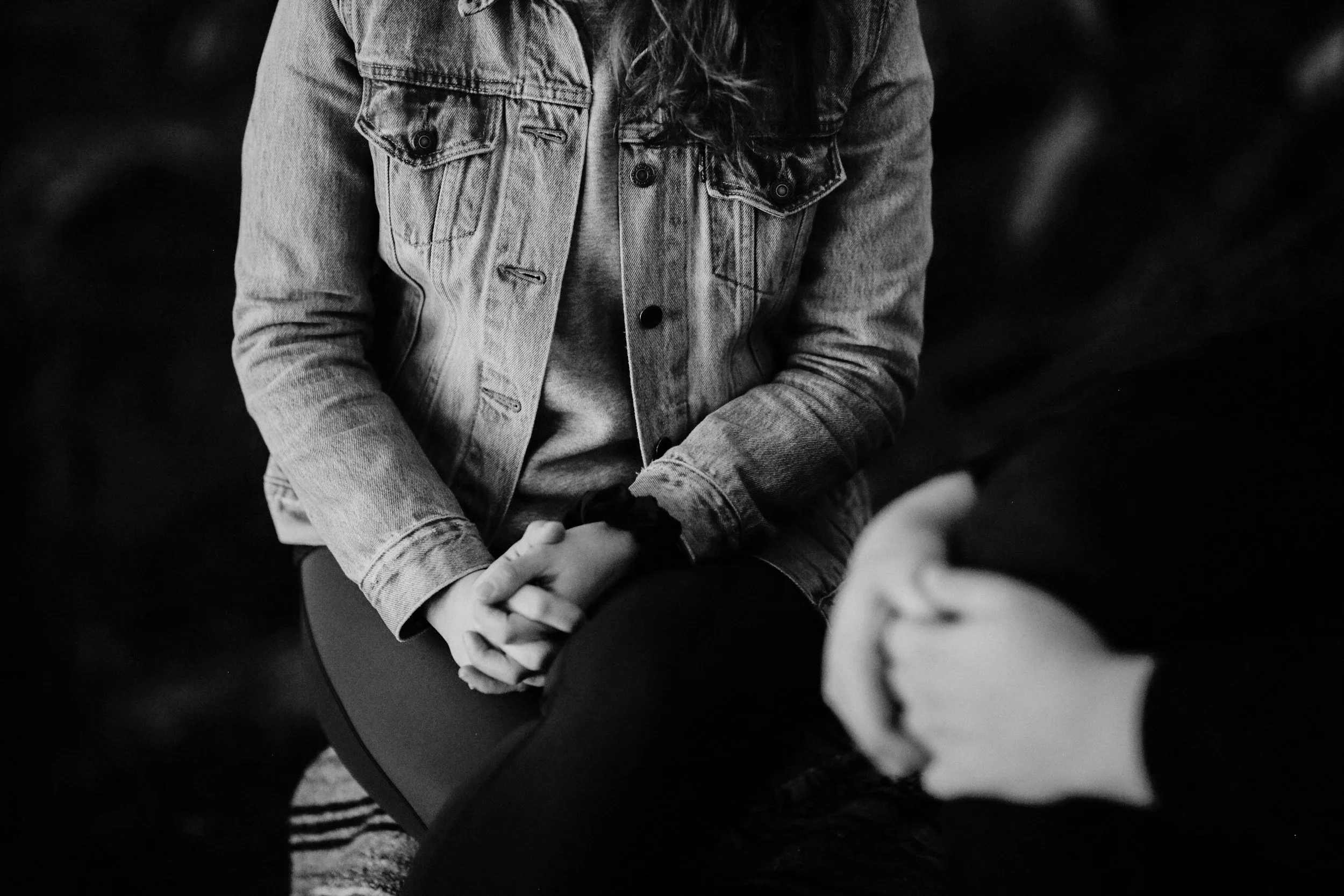 A person wearing a denim jacket with their hands clasped together in their lap, captured in black and white.
