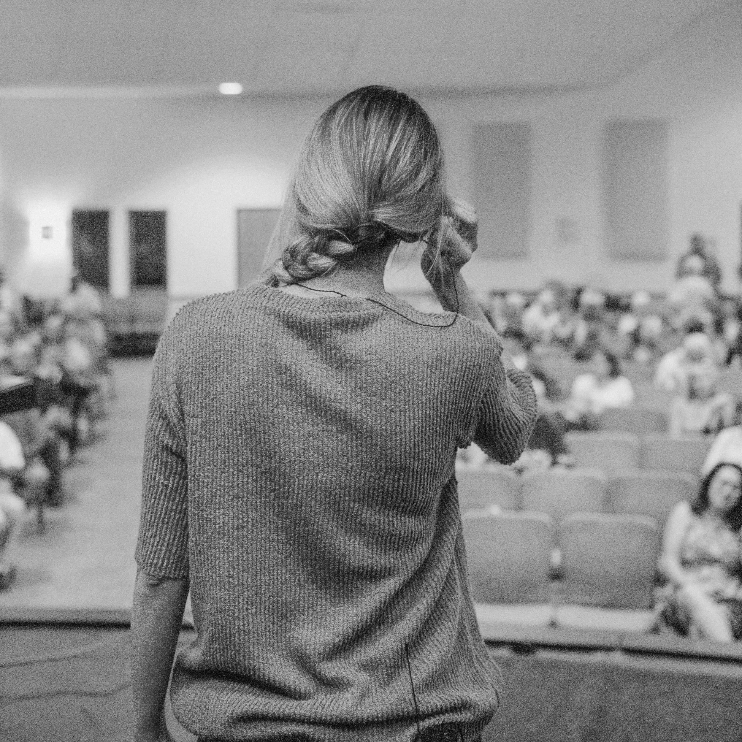 A woman with long hair in a braid wearing a textured sweater, seen from behind, standing in an indoor space with blurred background objects.