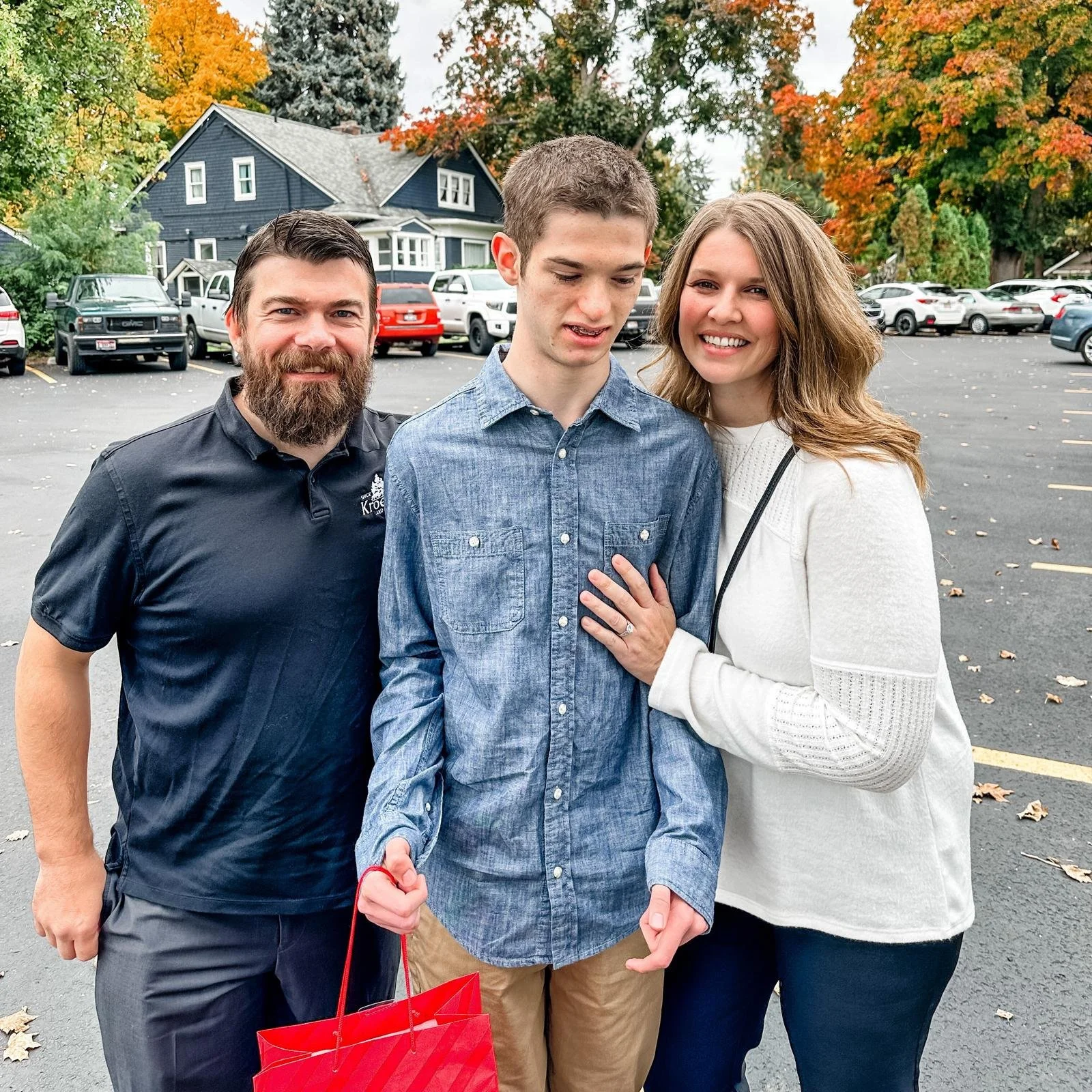 Three people posing for a photo in a parking lot with autumn trees in the background. The person in the center is holding a red gift bag.
