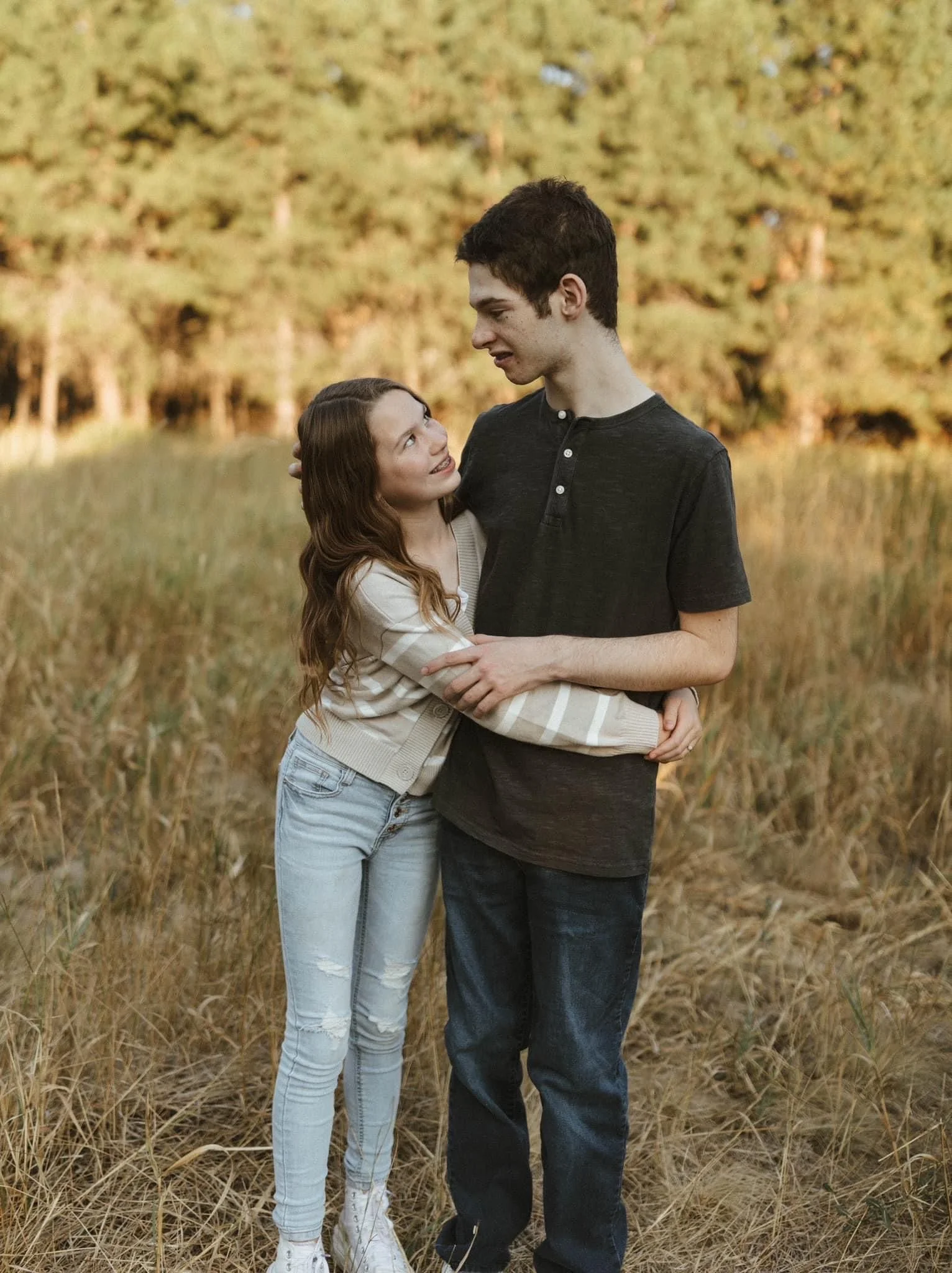 A young couple stands in a field, embracing and smiling at each other, with trees in the background.