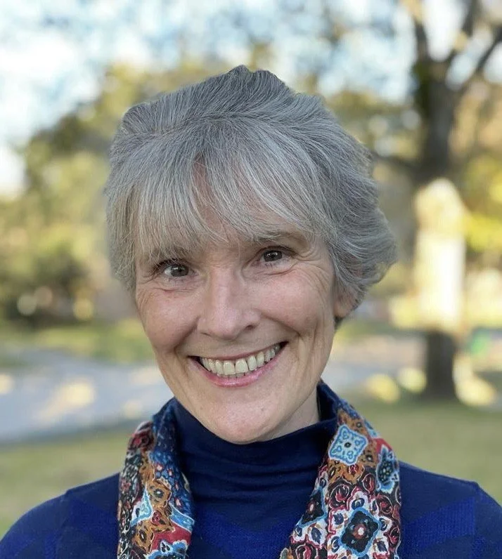 Older woman with gray hair smiling outdoors in natural light, wearing a colorful scarf and blue shirt.