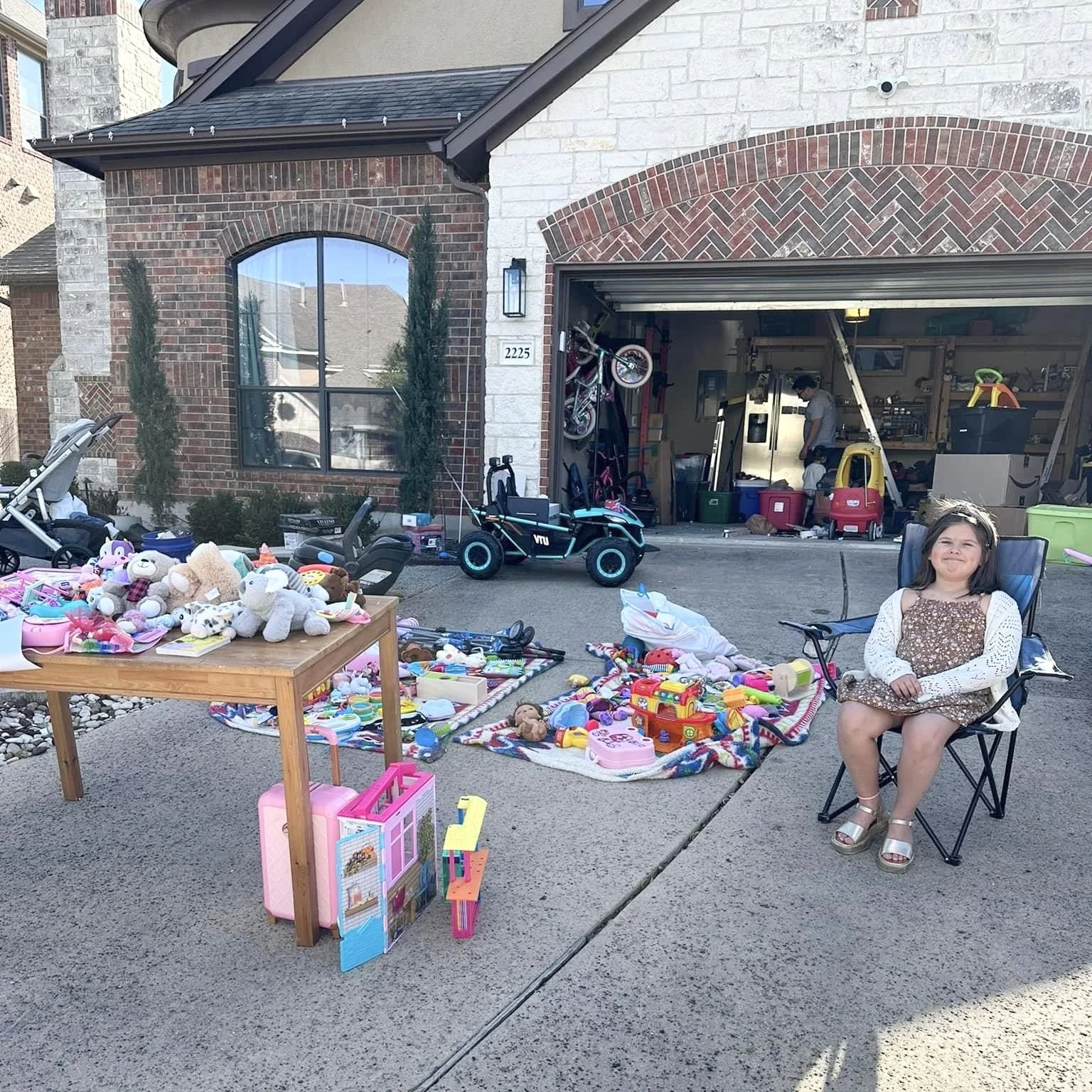 Garage sale with toys and stuffed animals on display, a child sitting in a camping chair, and a garage with bicycles and storage bins in the background.
