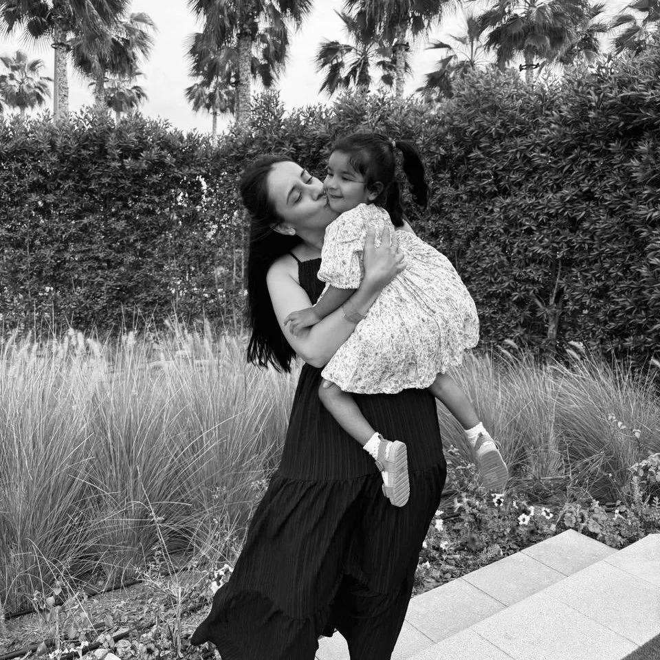 Black and white photo of a woman hugging a small child outdoors, surrounded by grass and palm trees in the background.