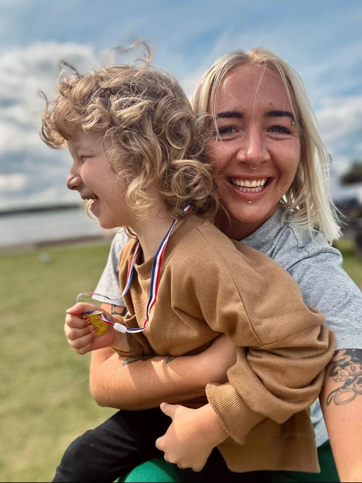 A happy moment with a child and an adult embracing outdoors. The child is wearing a medal around their neck, while both are smiling under a partly cloudy sky.