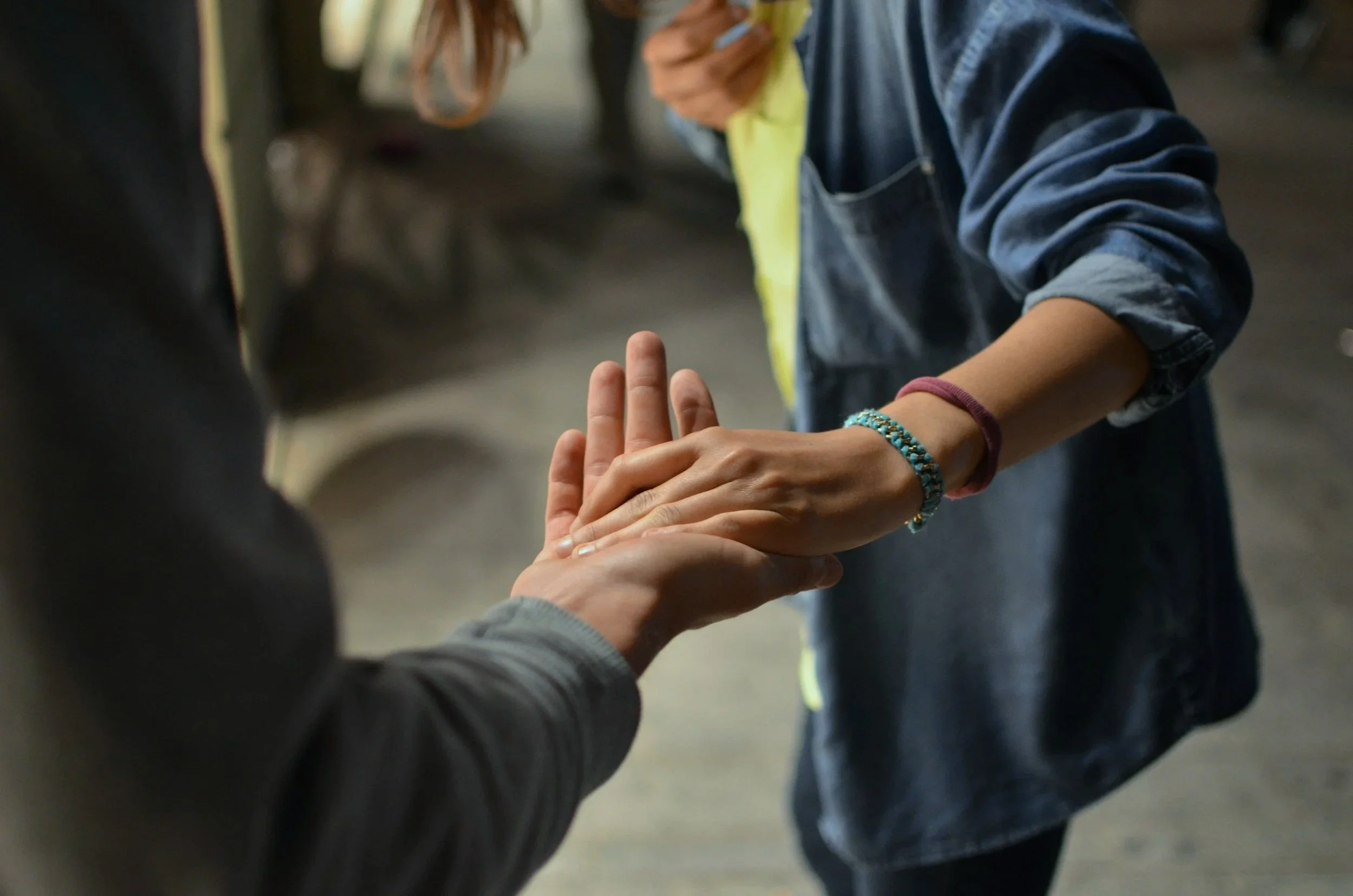 Two people holding hands, with one person gently grasping the other's hand and the other person's hand resting on top, in an outdoor setting.