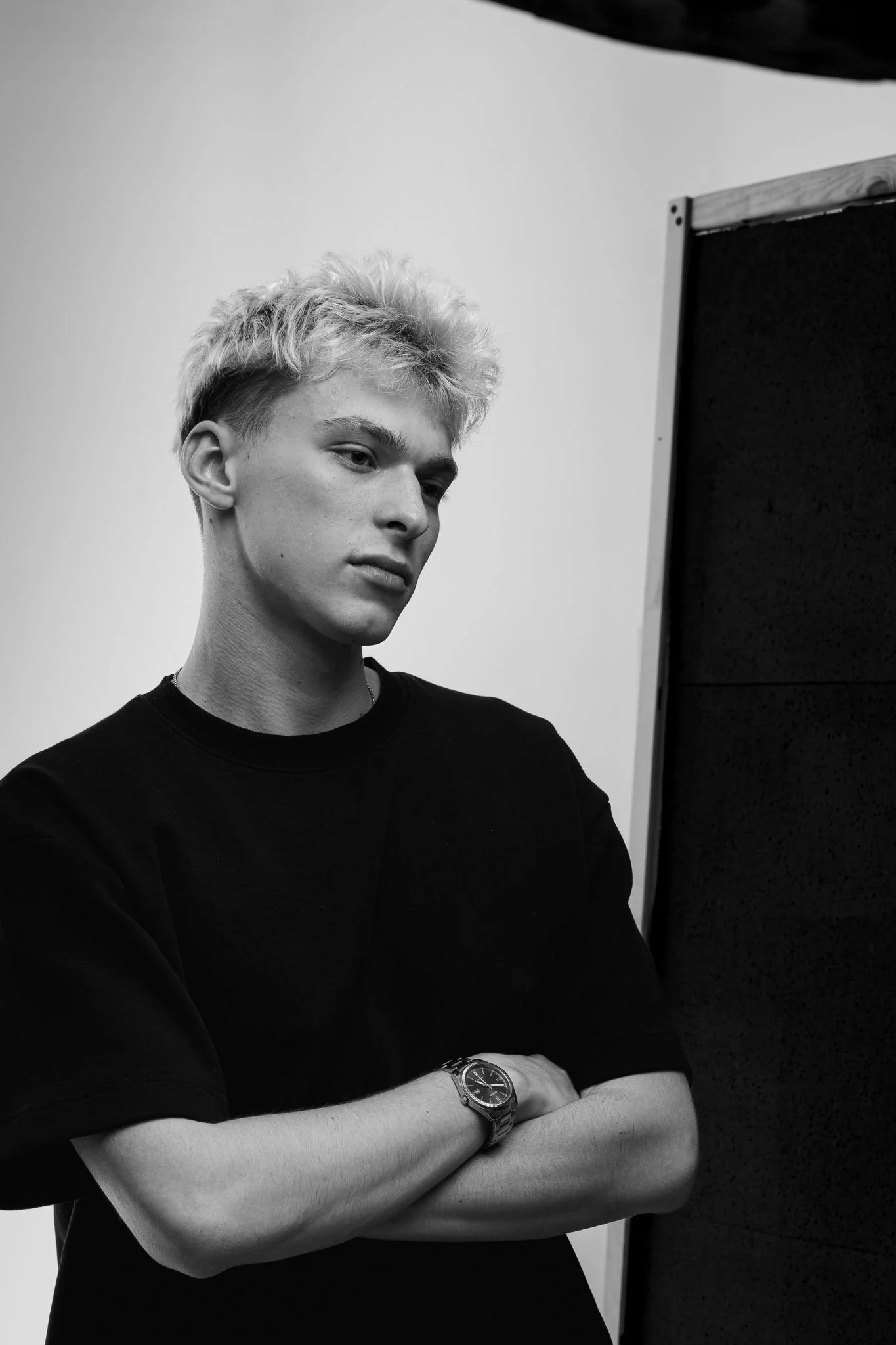 Black and white photo of a young man with a watch on his left arm, wearing a black shirt, standing with arms crossed in front of a dark partition, looking down with a serious expression.
