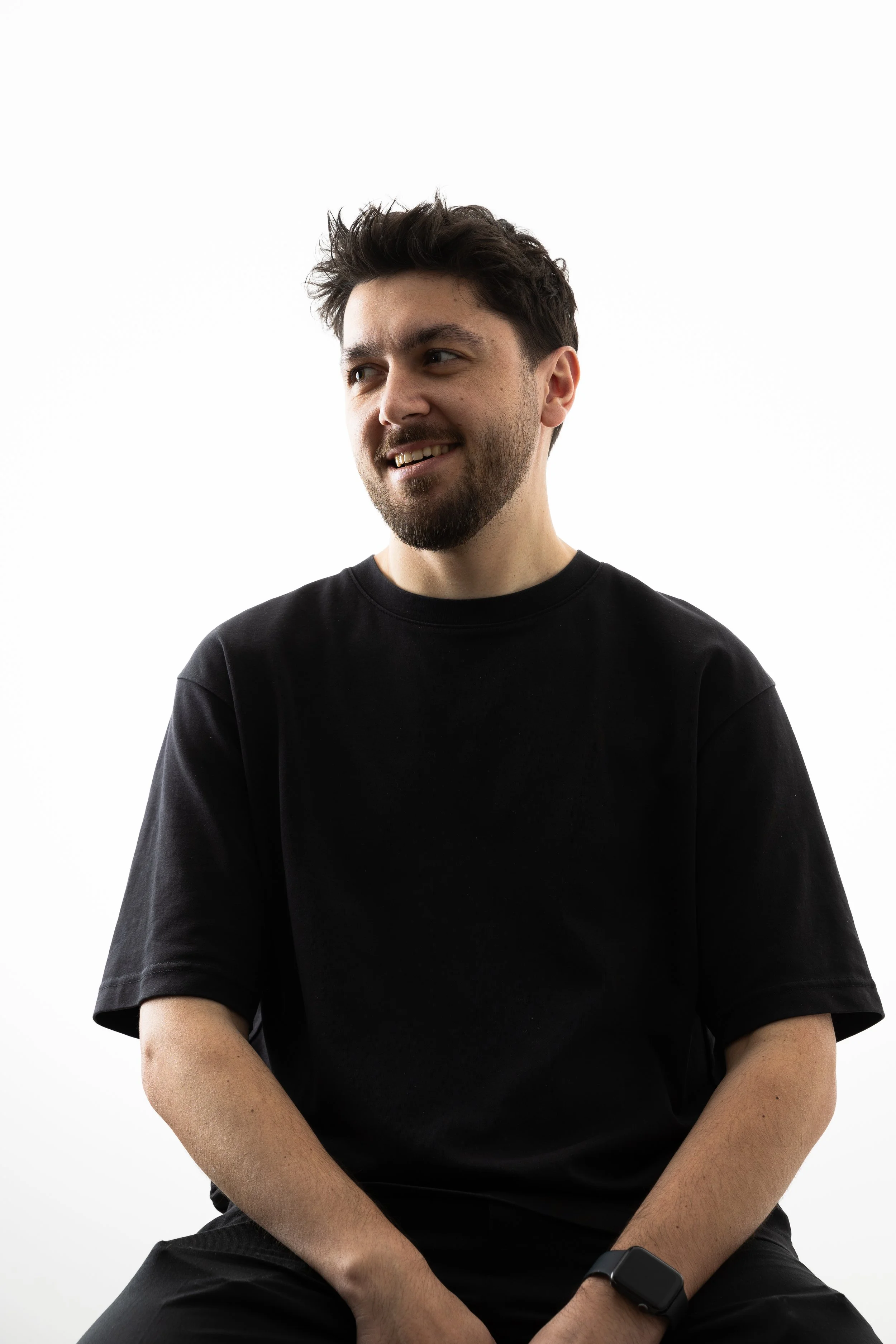 A young man with dark hair and a beard, wearing a black t-shirt and a smartwatch, is smiling and looking to his left against a white background.
