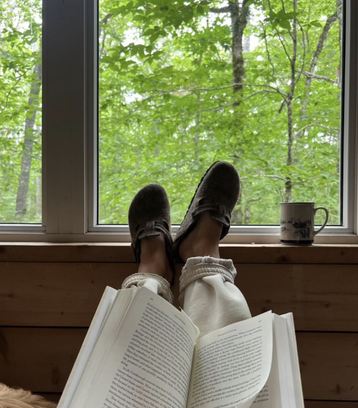 Person relaxing by a window with green trees outside, wearing slippers and reading a book, with a mug on the windowsill.