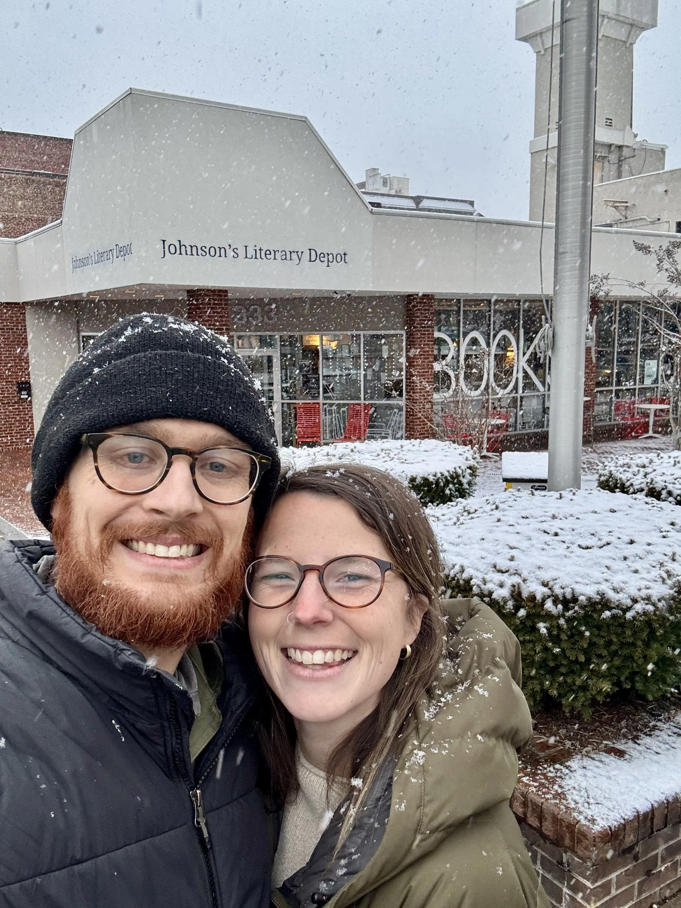 A smiling man and woman taking a selfie outside Johnson's Literary Depot on a snowy day.