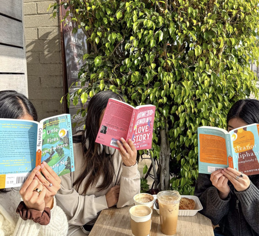 Three women sitting at an outdoor cafe table reading books with drinks and snacks, against a backdrop of a green shrub and a building wall.