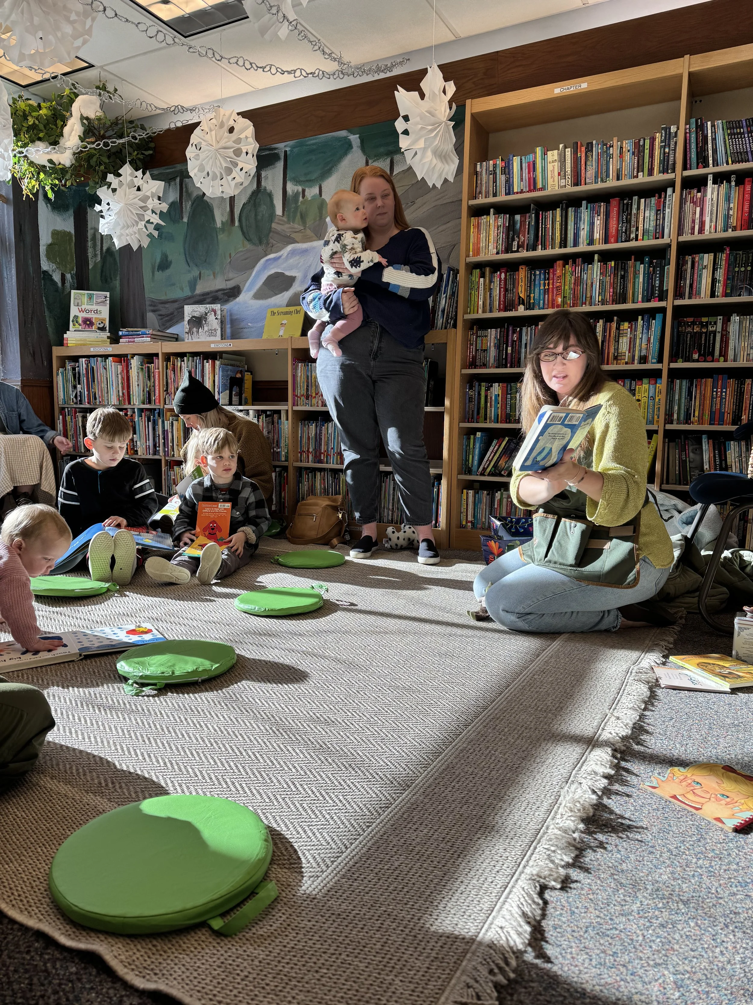 Children and adults participating in a storytime reading session in a library. Kids are sitting on a rug with green cushions, some reading books. Two women are reading, one is kneeling and the other is holding a child while standing.