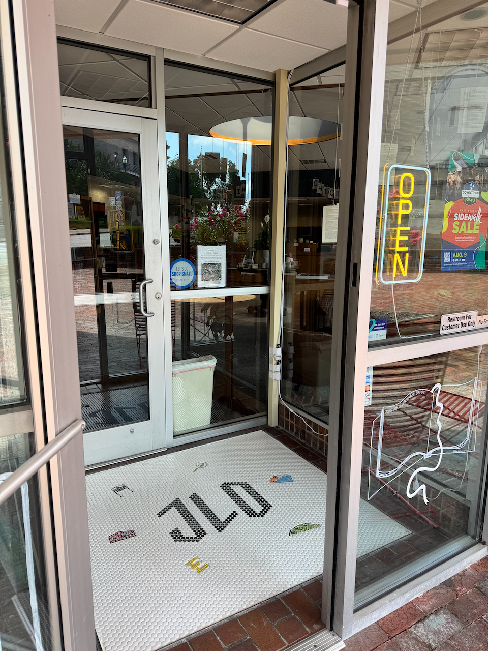 view of the entrance to a small shop or cafe through glass doors with an illuminated open sign, a "Shop Small" sign on the door, and a doormat with the word "SHOP" written upside down.