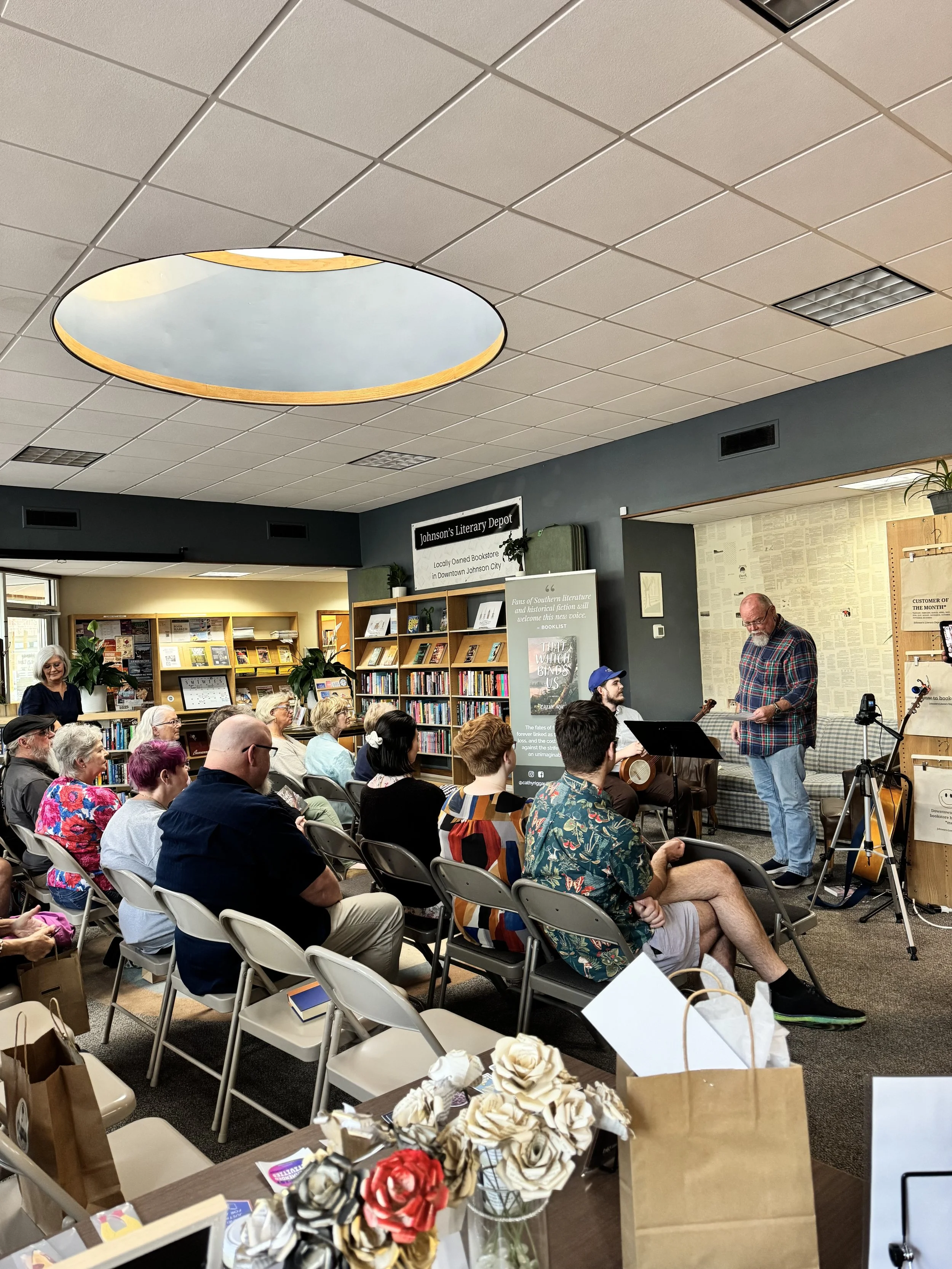 A man with a guitar and a man with a paper, standing in front of an audience at a bookstore event. Audience seated, some taking notes, others listening or watching. Bookstore shelves in the background, sign reading 'Johnson's Literary Depot'.