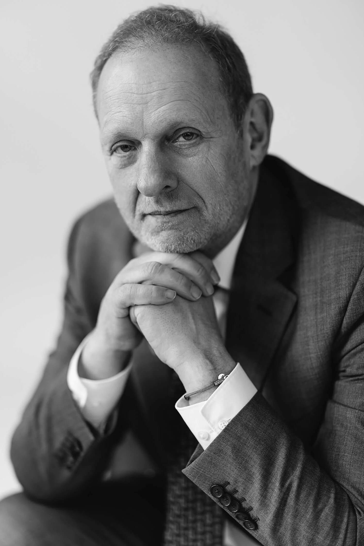 Zwarte en witte portretfoto van een oudere man in een grijze pak, met zijn handen onder zijn kin, naar de camera kijkend.