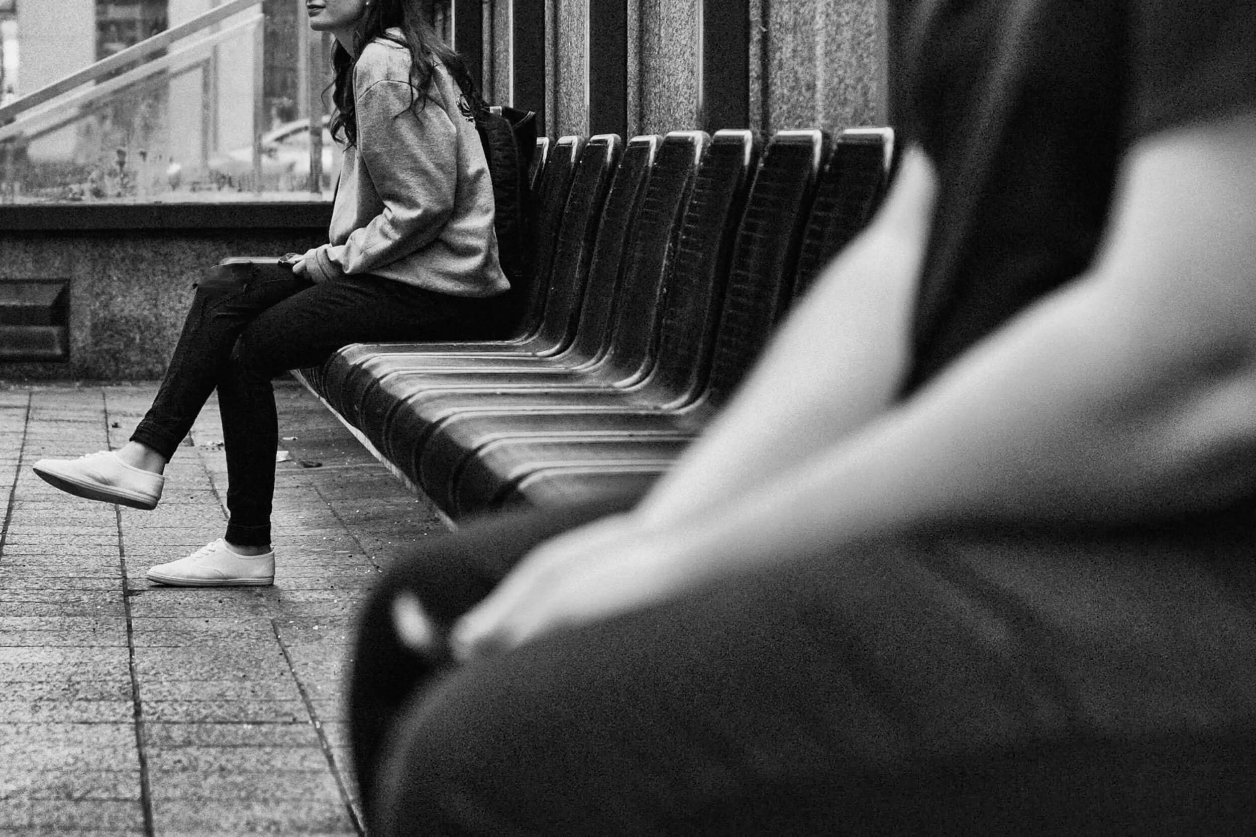 Een zwart-wit foto van een vrouw die op een bank zit in een station of bushokje, terwijl een andere persoon aan de rechterkant uit de lens halve of niet zichtbaar is.