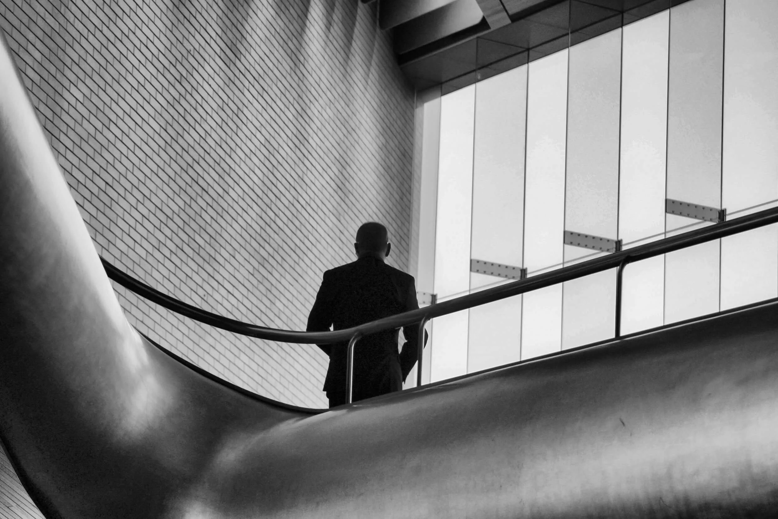 Silhouet van een man in een pak die naar binnen kijkt, staand op een trap in een modern gebouw met grote ramen en een bakstenen muur, zwart-wit foto.