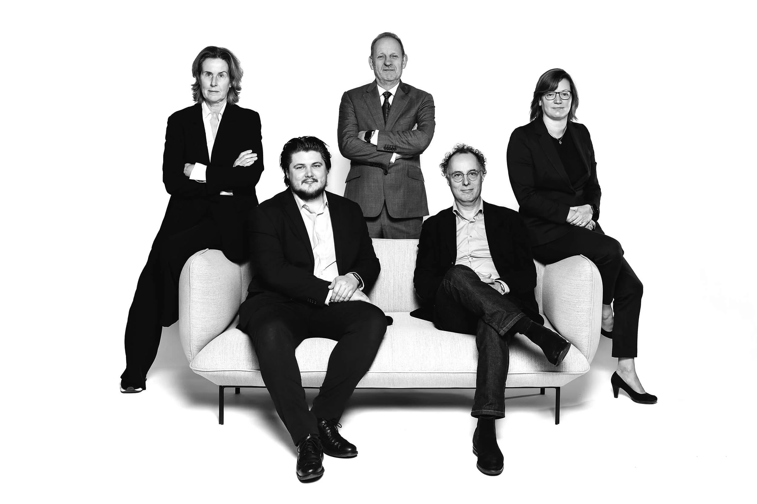 Zes mensen, drie mannen en drie vrouwen, poseert in professionele kleding tegen een witte achtergrond. Drie mensen zitten op een bank en de andere staan erachter.