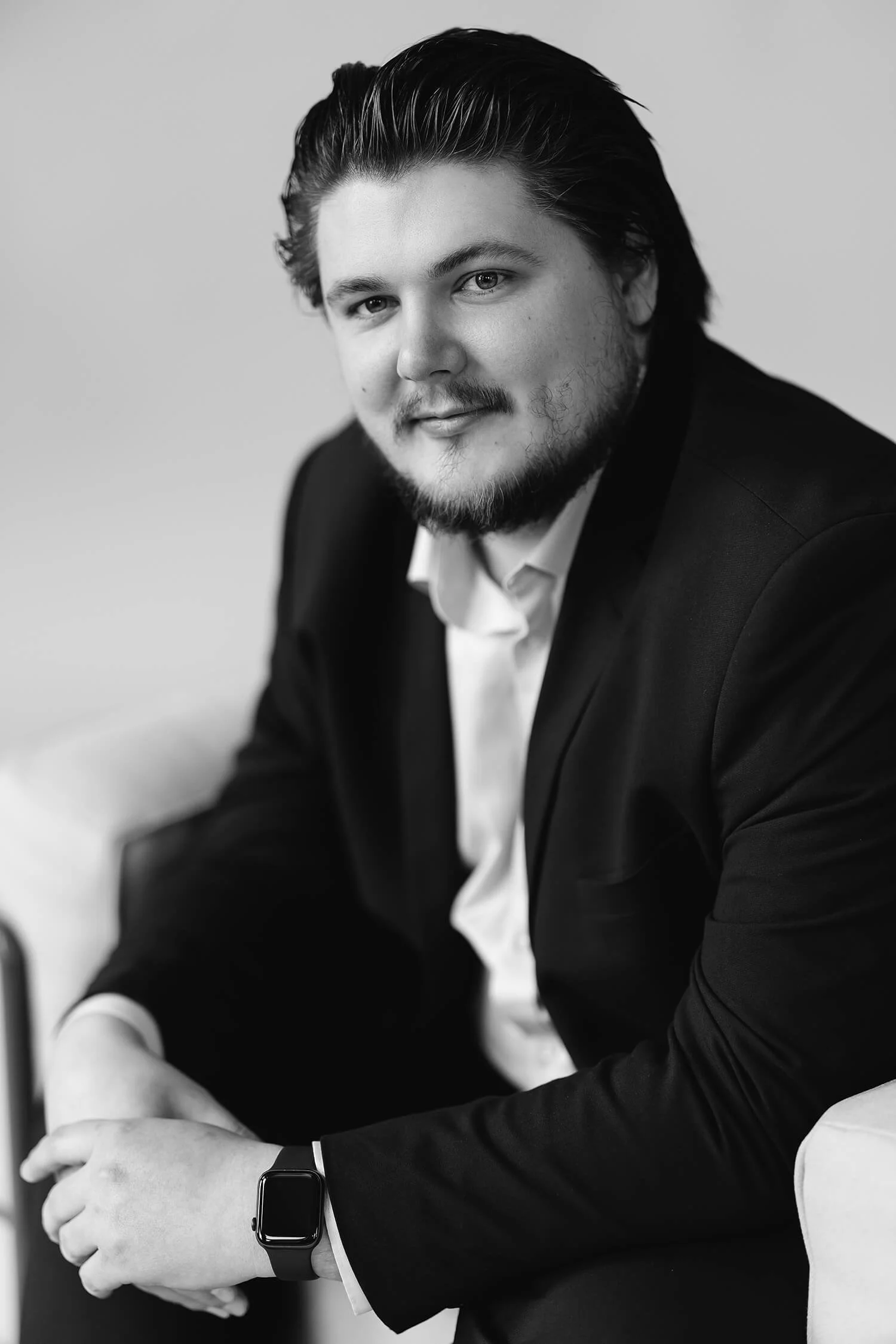 Zwart-witte portretfoto van een man met donkere geverfde haar, een baard en een glimlach, gekleed in een zwarte jas en een witte overhemd, zittend met een smartwatch op zijn pols.