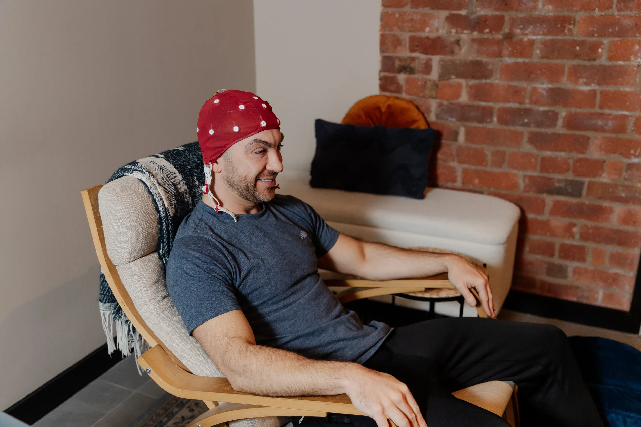 A man with a red swim cap with white polka dots, wearing a navy blue t-shirt, sitting on a wooden chair with a cushioned seat and backrest, smiling, against a brick wall background.