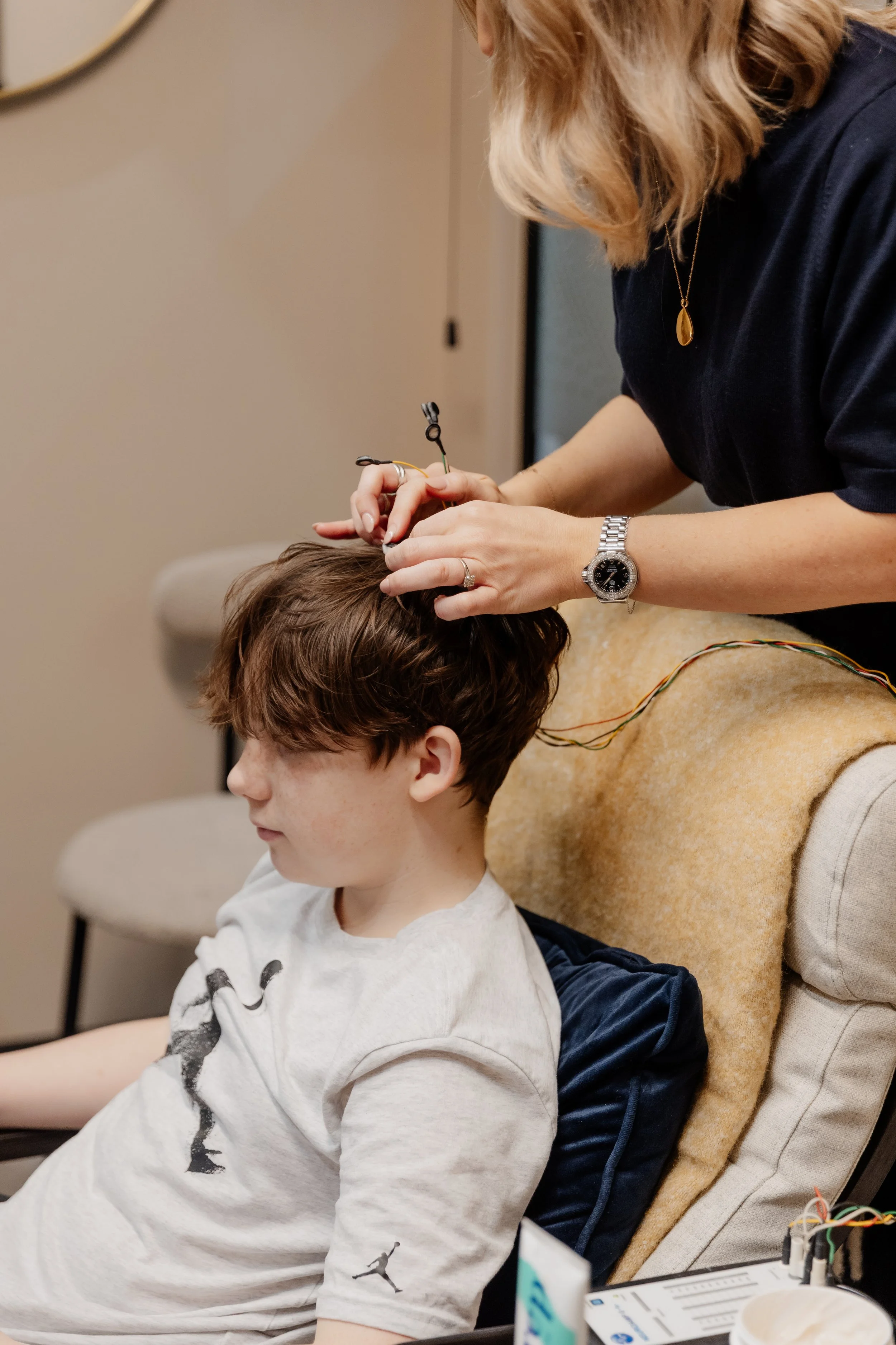 A woman is giving a young boy an EEG or medical test by placing electrodes and wires on his head in a clinical setting.